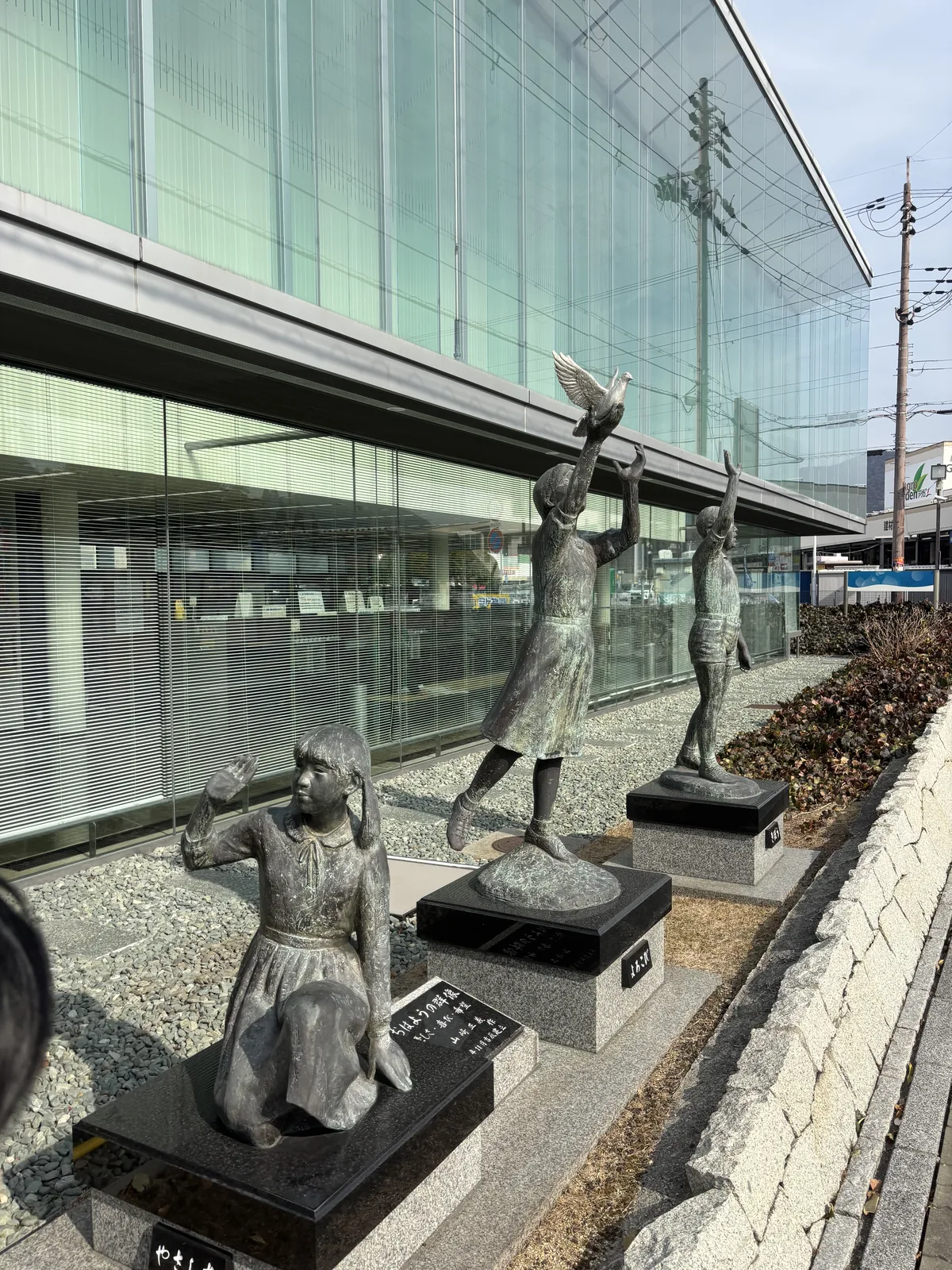 赤穗市立圖書館的外觀，兩層樓高的玻璃立面沿街延伸，立面之前的碎石植栽上立著三尊銅像，分別是坐著揮手的少女、放飛鴿子的少女與呼喊的男孩，右側是電線桿與晴空