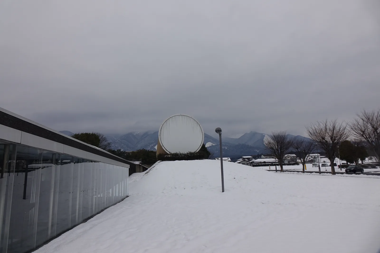 奈義町現代美術館的外觀，前景是覆蓋積雪的草坡，坡頂上露出一個橫躺的銀色圓筒，圓形的端面朝向畫面，左側是一道低矮的玻璃迴廊，背景是灰色的天空與積雪的山稜