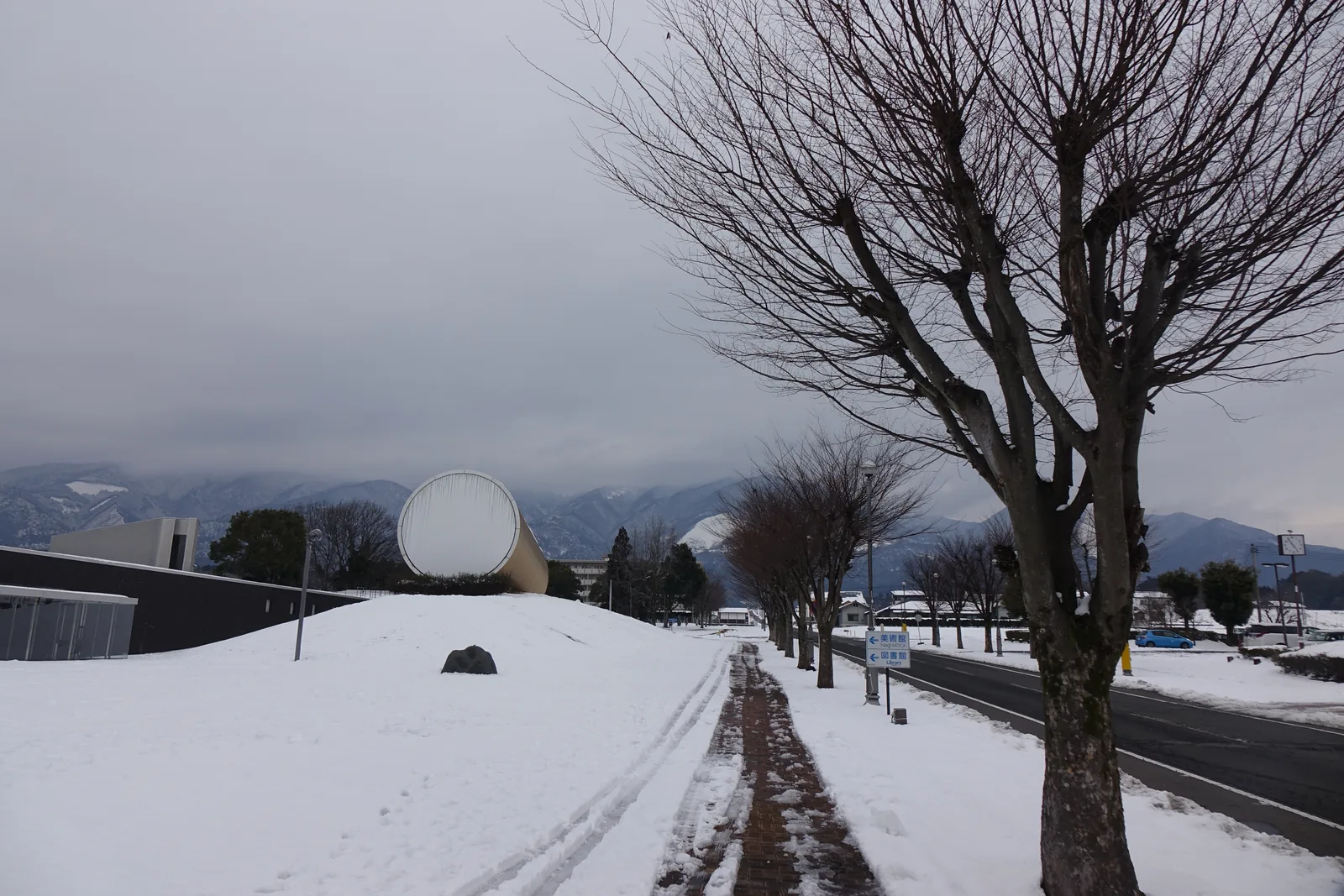 館前的道路，右側是一排落葉的行道樹，左側的人行道上堆著積雪，道路沿線可見美術館的銀色圓筒與低矮的量體，背景是被雲遮住的山稜