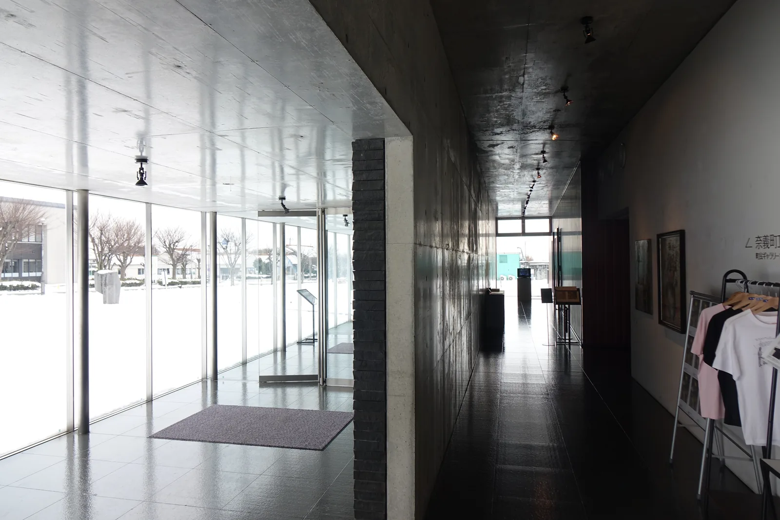 入口內的 lobby，左側是整面的落地玻璃與門，玻璃外是積雪的廣場，中央是一根深灰色砌體的牆柱，右側是一條天花低暗的長通道，通道盡頭是另一面玻璃，牆邊掛著 T 恤