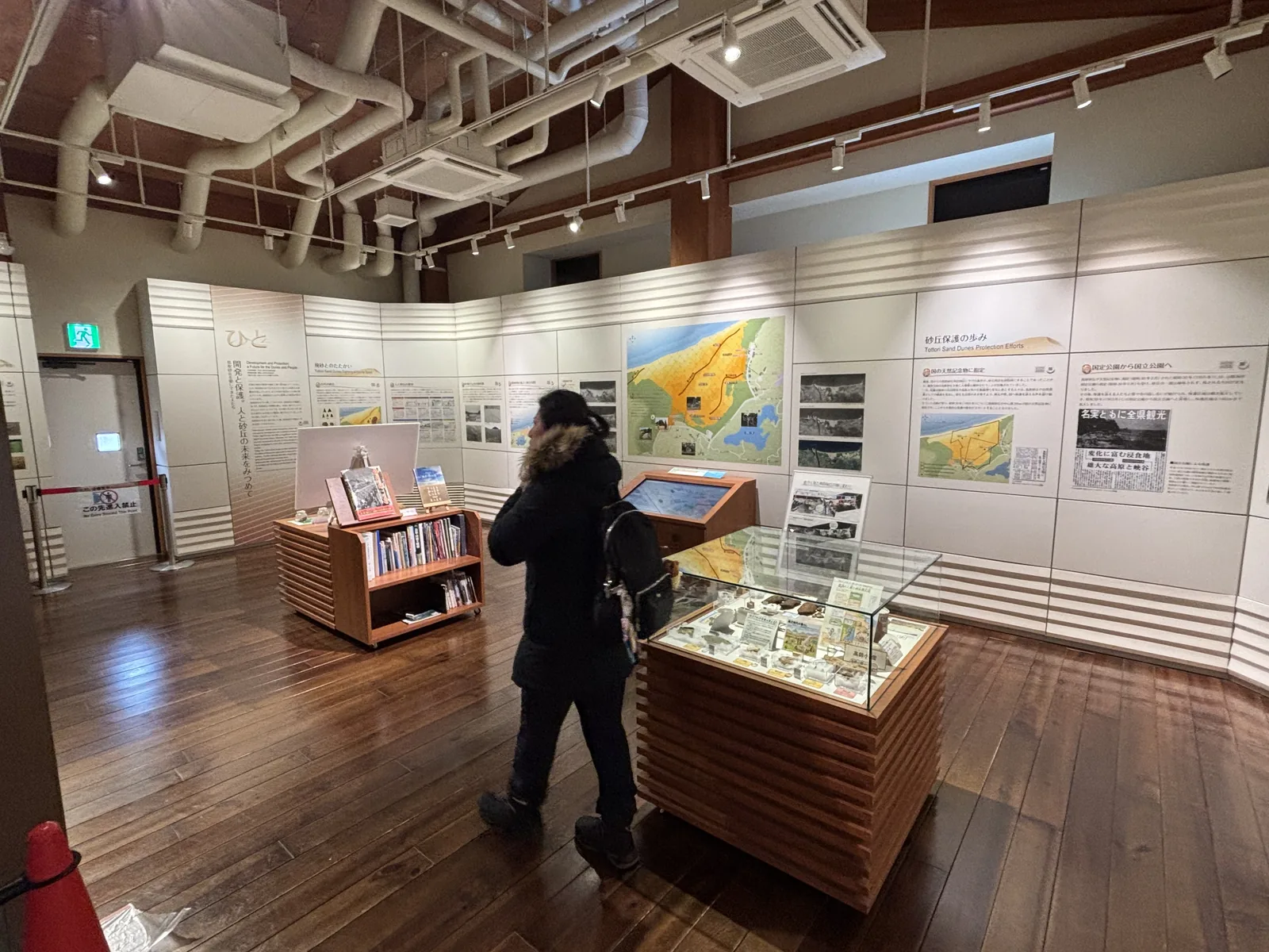 遊客中心的展示室，地面是深色的木質地板，四周是白色的展示牆，牆上貼著砂丘的地圖與照片，中央是木質的展示 case 與書架，天花上露出木梁、空調管線與軌道照明，一位參觀者背向鏡頭站在展示 case 旁