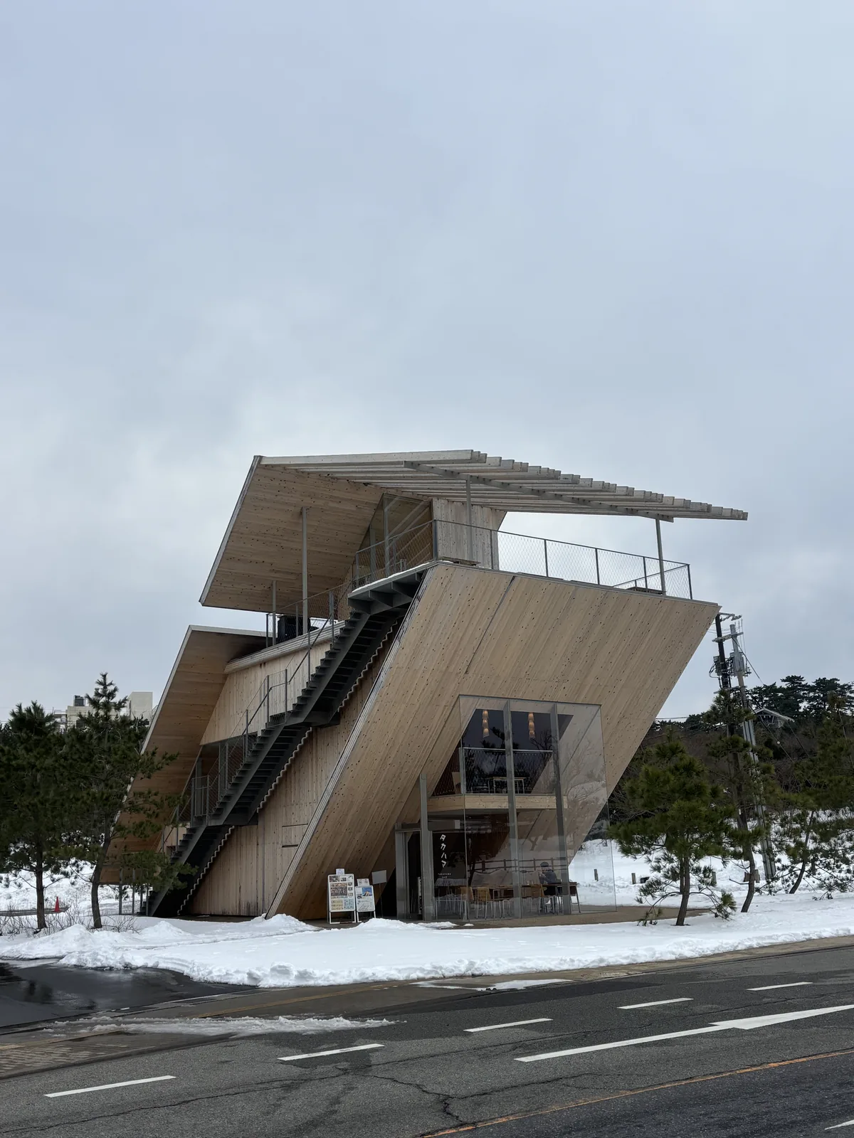Takahama Cafe 的外觀，一座木構的量體向上傾斜，外側沿斜面設一道鋼構的外部階段，頂端是一片以細長構件組成的遮陽屋頂，前方是道路與積雪的草地，兩側是松樹