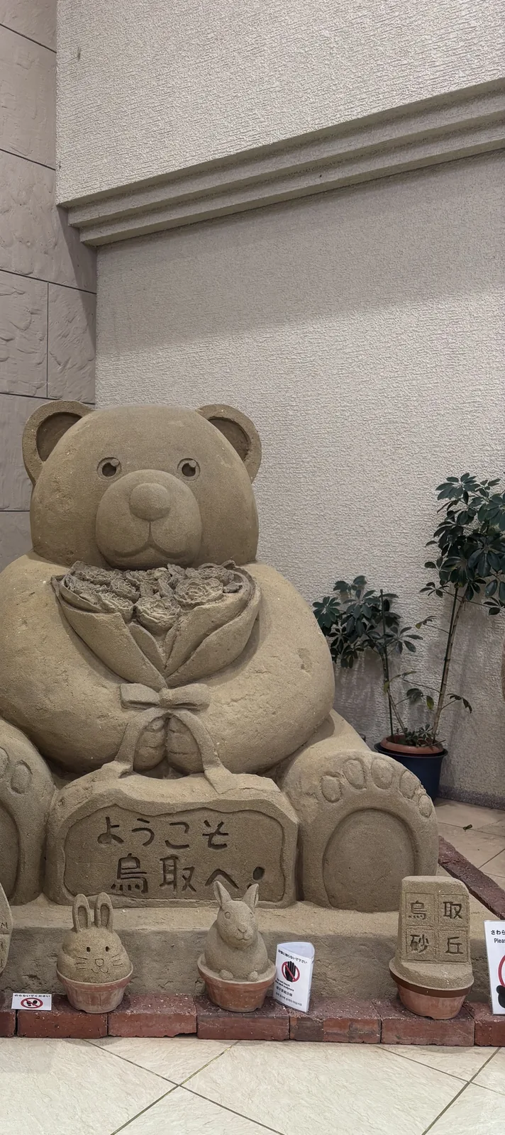 館內一角的砂像，一隻以砂堆塑的熊坐在磚砌的台座上，胸前捧著一束砂做的花，台座正面刻著歡迎來到鳥取的字樣，前方擺著兩隻砂做的兔子與一塊刻有鳥取砂丘四字的砂塊，背後是帶水平線腳的米色塗裝牆，右側是盆栽植物，地面是淺色地磚