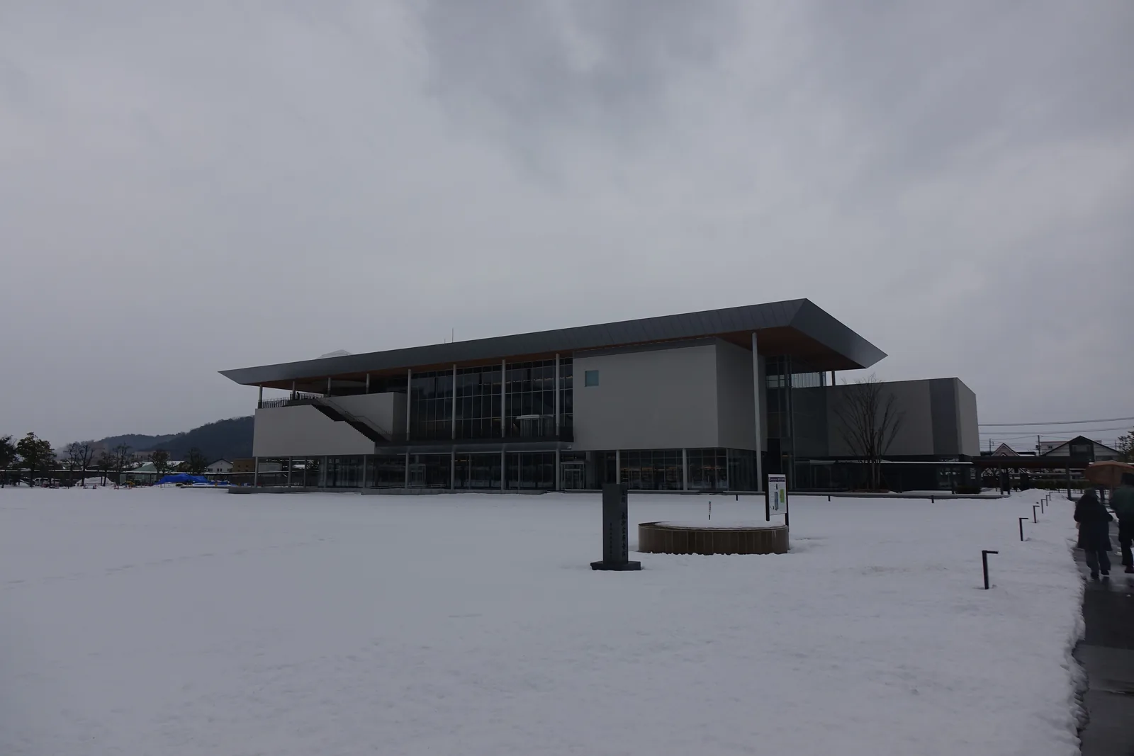 自南側的史跡公園走向美術館，前景是覆雪的廣場與一條除過雪的步道，廣場上立著幾根黑色的低矮柱樁，遠景是美術館低平的量體與大屋頂，背後是被雪覆蓋的山