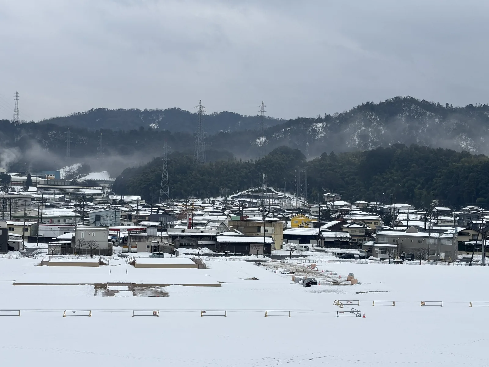 自三樓展望露台望向南側的史跡公園，覆雪的地面上以低矮的木柵與標示排出遺構的範圍，遠景是倉吉市街的屋頂與一道籠罩在雲霧中的山稜