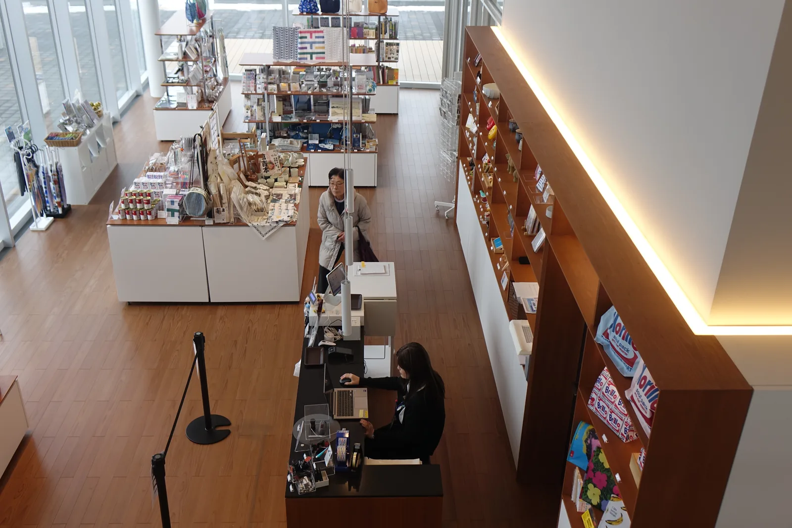 自二樓俯看一樓的 museum shop，木地板上排著白色的展示台與貨架，貨架上陳列著各色商品，中央是一座黑色的收銀台，右側是一道木色的牆與間接照明