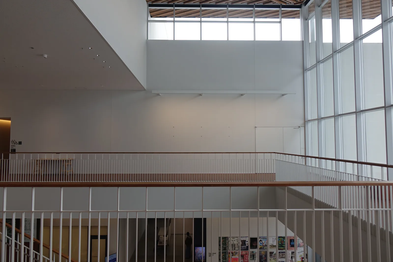 自二樓望向挑空另一側的 gallery，白色的欄杆沿著挑空延伸，對面是三樓的走廊與高窗，下方一樓的牆上貼著海報，右側是整面的玻璃立面
