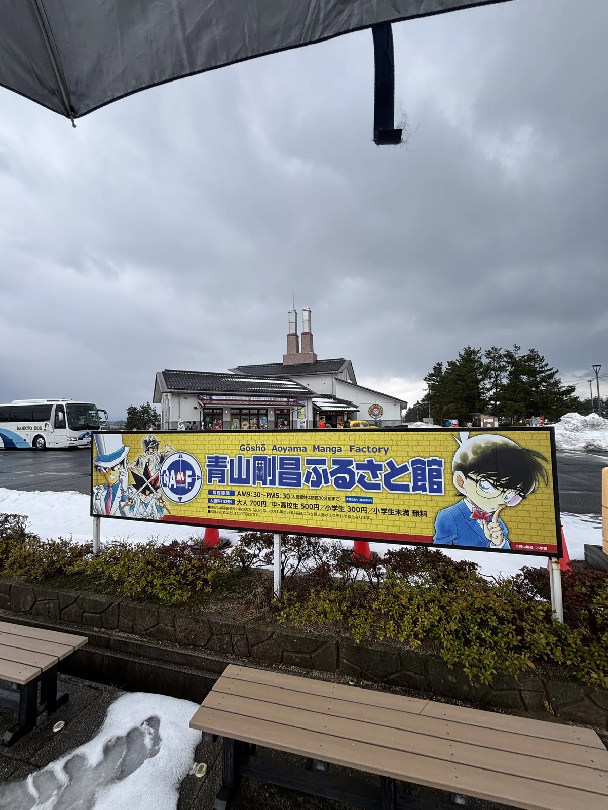 故鄉館前的館名看板，看板上寫著館名與英文名 Gosho Aoyama Manga Factory，旁邊畫著柯南的插圖，看板之後是故鄉館的建築與屋頂上的兩支煙囪狀構件，前景是覆雪的植栽與木製長椅