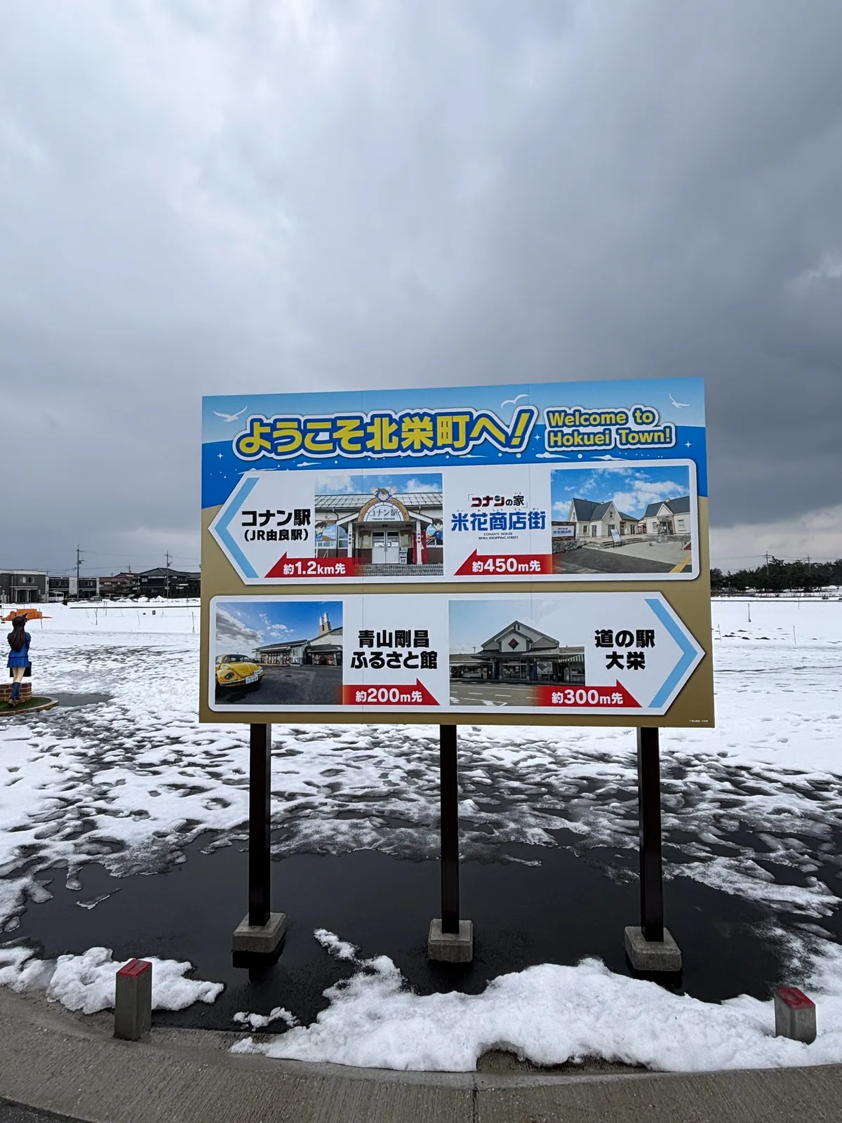 北榮町的歡迎看板，藍色的看板上以照片與距離標示柯南車站、米花商店街、青山剛昌故鄉館與大榮公路休息站四個地點，看板立在覆雪的空地上，背後是灰色的天空