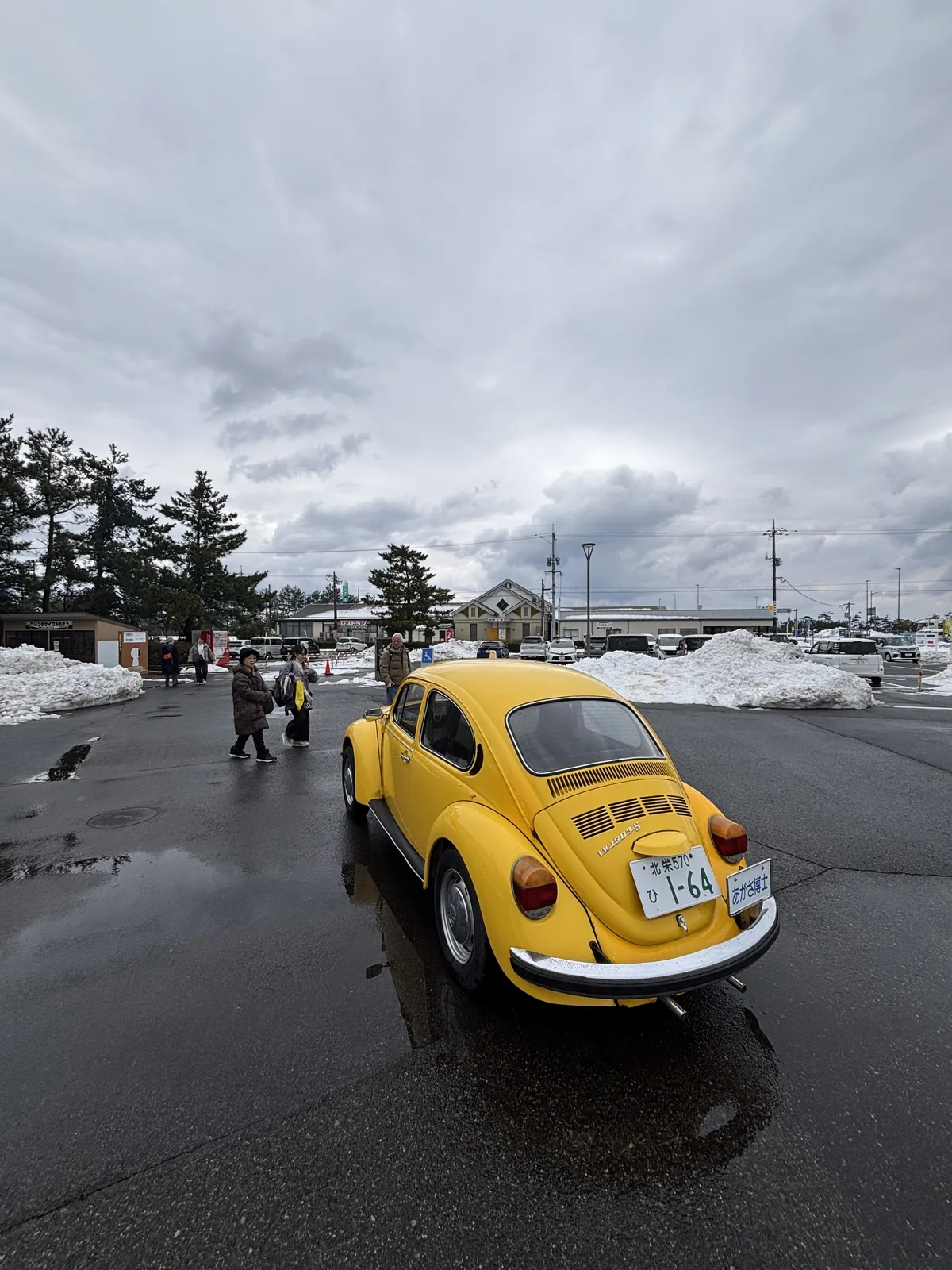 故鄉館駐車場上的一輛黃色 Volkswagen Beetle，自車尾斜後方拍攝，車牌號碼為 1-64，背景是濕的柏油路面、殘雪、幾位遊客與遠處的建築