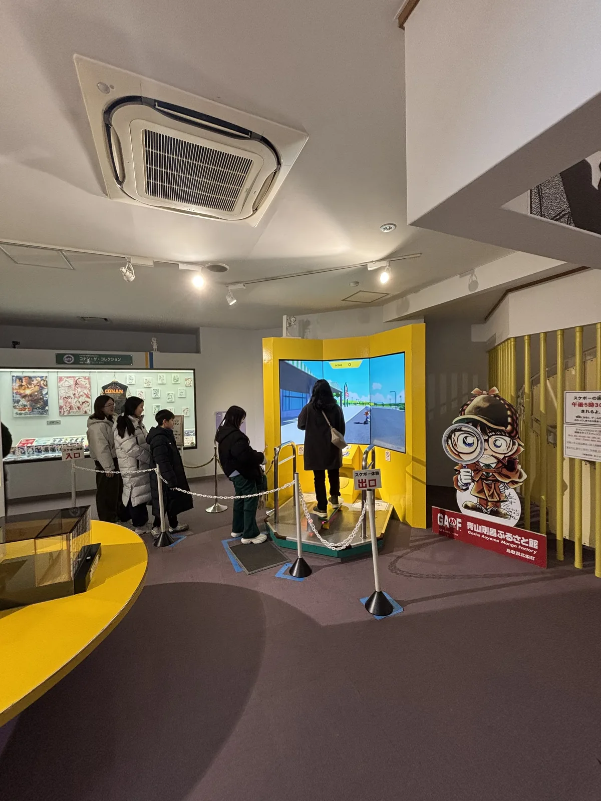 館內的一間展示室，白色的平天花上吊著空調機與軌道照明，地面是深紫色的地毯，中央是一組黃色的曲面隔板，隔板內有一面大螢幕，幾位參觀者排隊等候，左側沿牆是展示櫃與說明板，右側立著一座卡通人物的立牌
