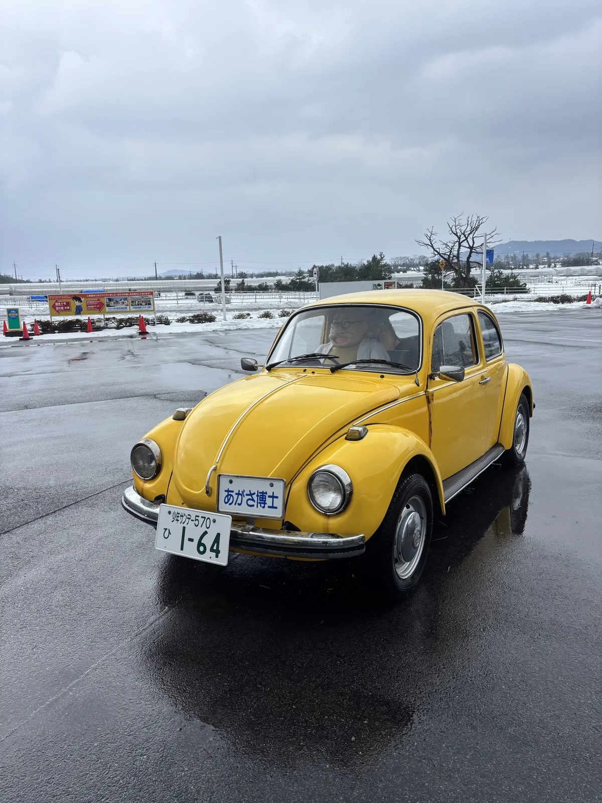 一輛黃色的 Volkswagen Beetle 停在故鄉館的駐車場上，自車頭斜前方拍攝，車牌號碼為 1-64，背景是覆雪的田地與遠山