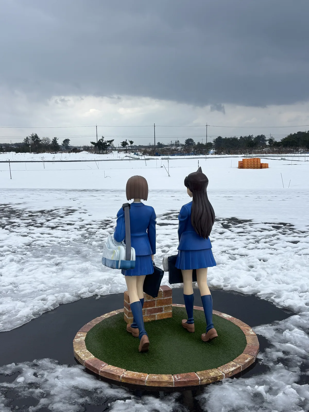 同一組彩色像的背面，兩位女學生背著書包，望向遠方覆雪的田地與道路，台座是圓形的人工草皮