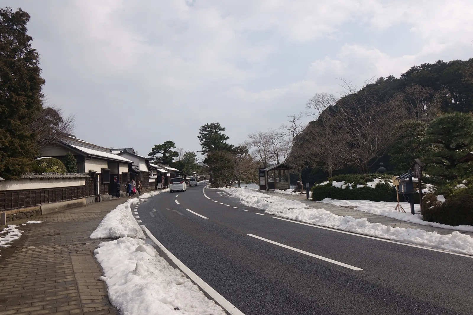 鹽見繩手的街景，左側是一列低矮的武家屋敷與土塀，屋頂殘留積雪，中央是向前延伸的柏油道路與堆在路緣的積雪，右側是內堀邊的松樹與城山的樹林，天空是陰天