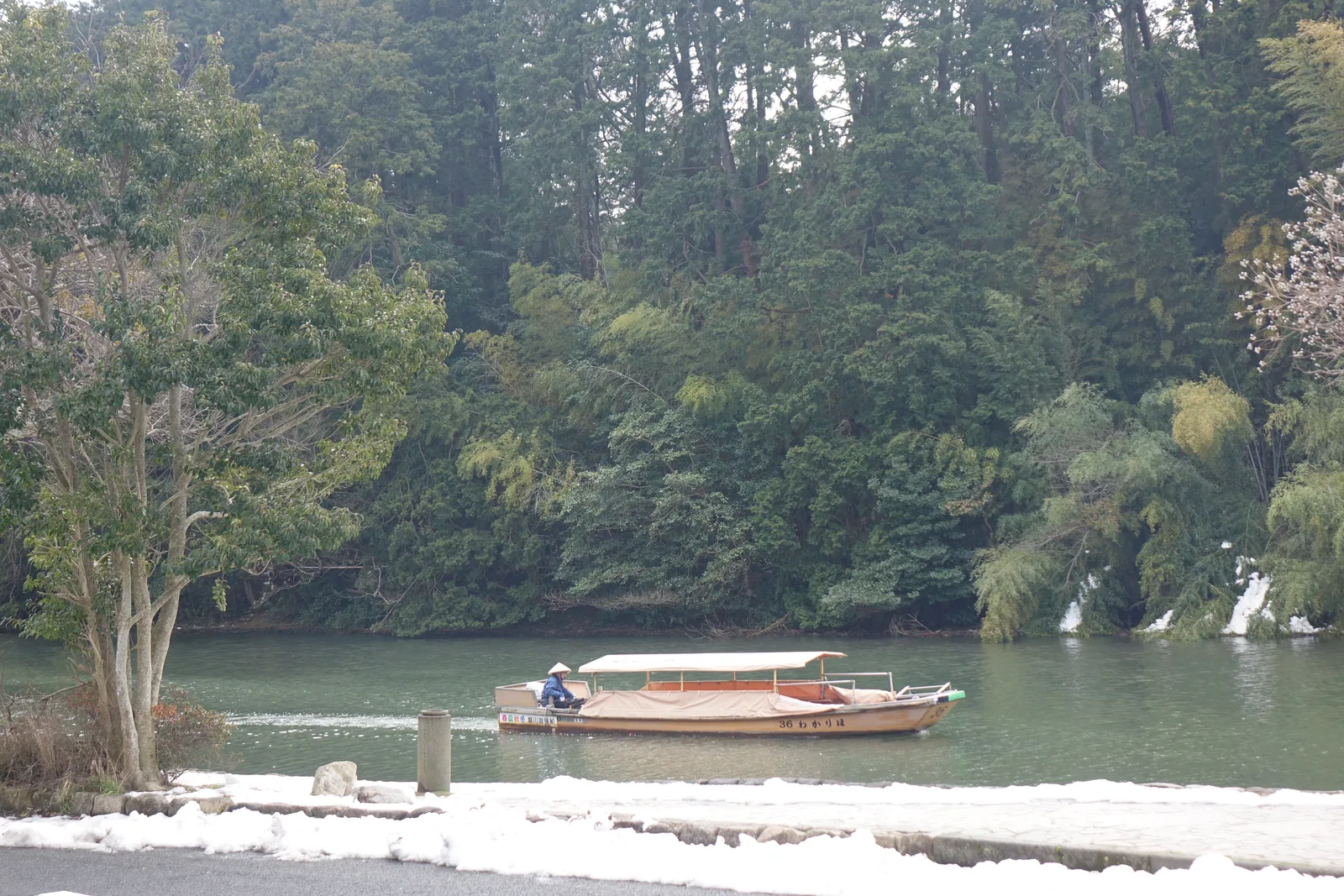 內堀的水面上，一艘有頂篷的遊覽船駛過，對岸是城山茂密的常綠樹林與竹叢，前景是覆雪的堀端