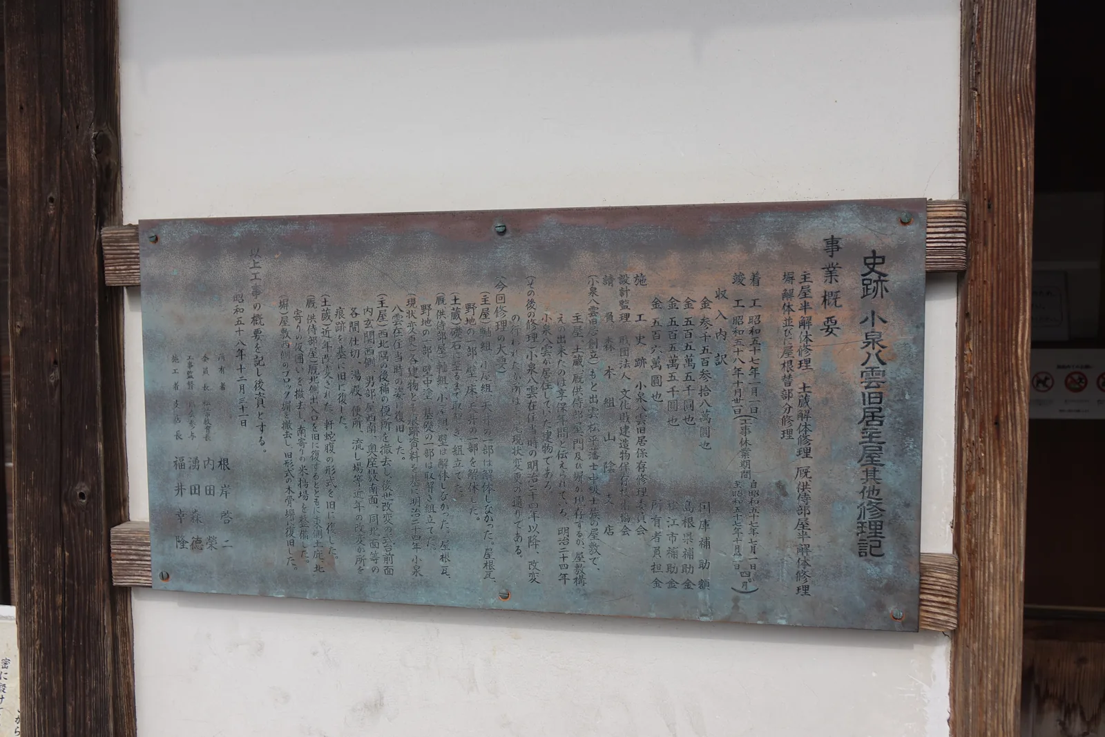 白色抹灰壁面上，以兩根木條固定一塊青銅色的銅牌，銅牌上直書「史跡小泉八雲舊居主屋其他修理記」與修理的經過，左側是一根深色的木柱