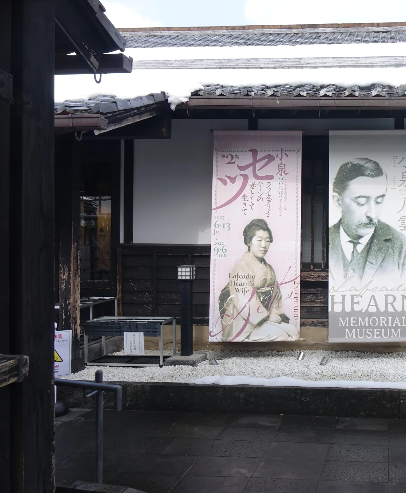 小泉八雲記念館的正面，一座木造平屋的和風建築，黑瓦屋頂上殘留積雪，白色壁面與深色木格子窗之前垂掛著兩幅布幕，一幅是 Setsu 的畫像，一幅是 Hearn 的肖像與英文館名