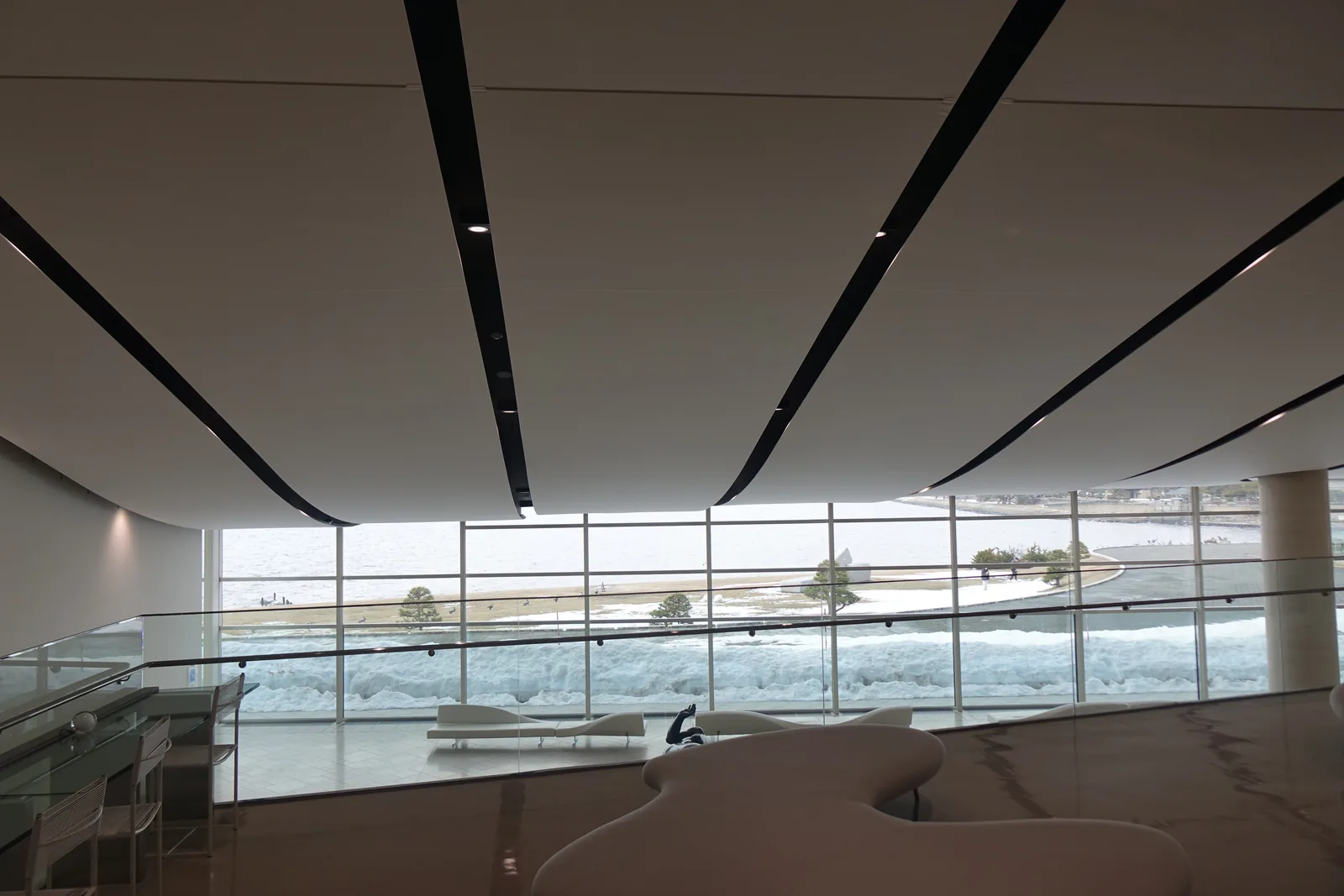 二樓 art library 前方，越過玻璃欄杆望向一樓大廳與湖面，前景是白色的曲線形長椅，中景是欄杆旁的閱覽桌，遠景是整排落地玻璃與覆雪的湖岸，天花上數道暗色的線條斜向延伸