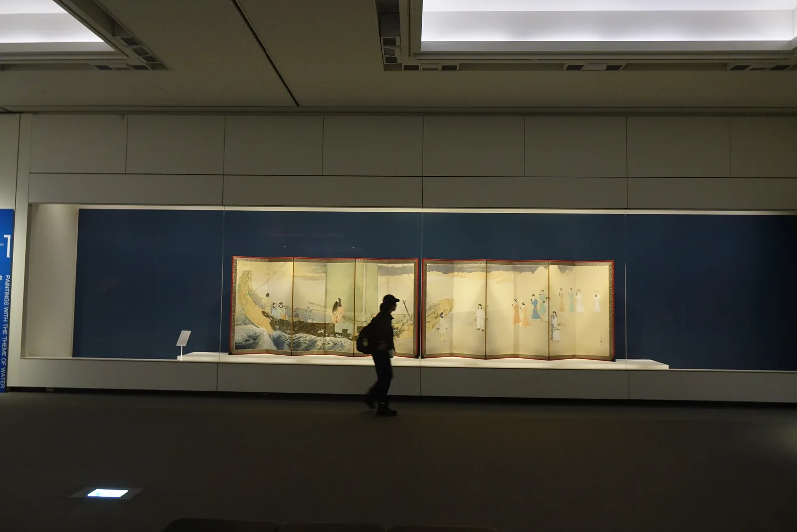 展示室內部，深藍色的展示牆前方是兩件並列的金地屏風，位於一道長條的玻璃展示櫃內，一位參觀者站在屏風前，天花較低並有間接照明，地面昏暗