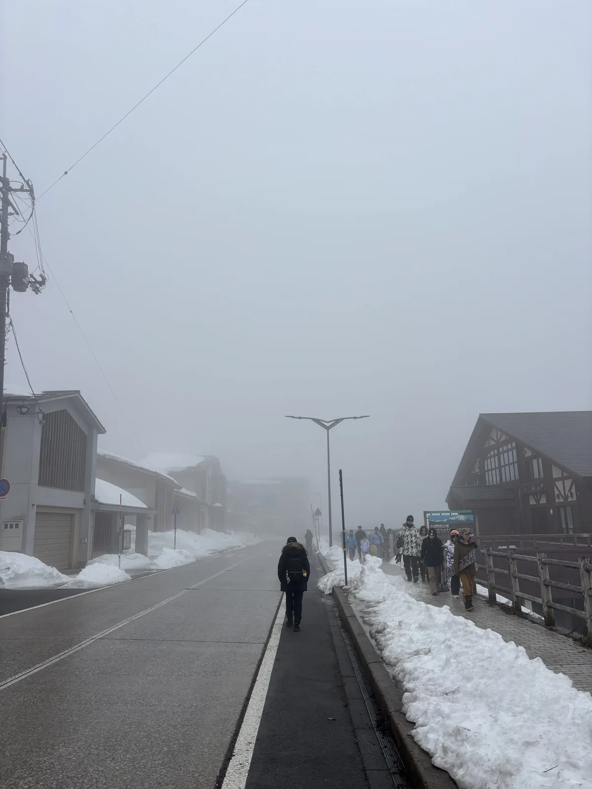 濃霧中的道路，左側是車道與路緣，右側是人行道上的行人與覆雪的路肩，遠處右側是國立公園中心深色木構的山牆，道路與建物都隱沒在霧裡