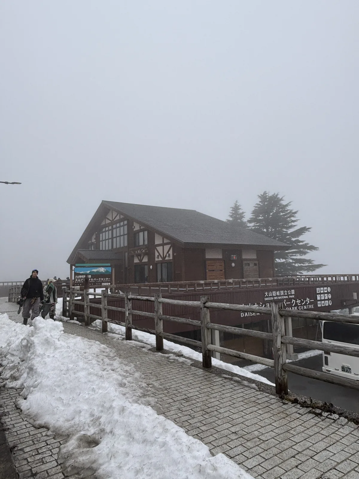 自縣道側望國立公園中心，前景是覆雪的人行道與木製的欄杆，中央是一棟二層建物，上層為深色木構的山牆與格子窗，下方延伸出一圈平台，平台邊緣掛著館名看板，左側有兩名步行者
