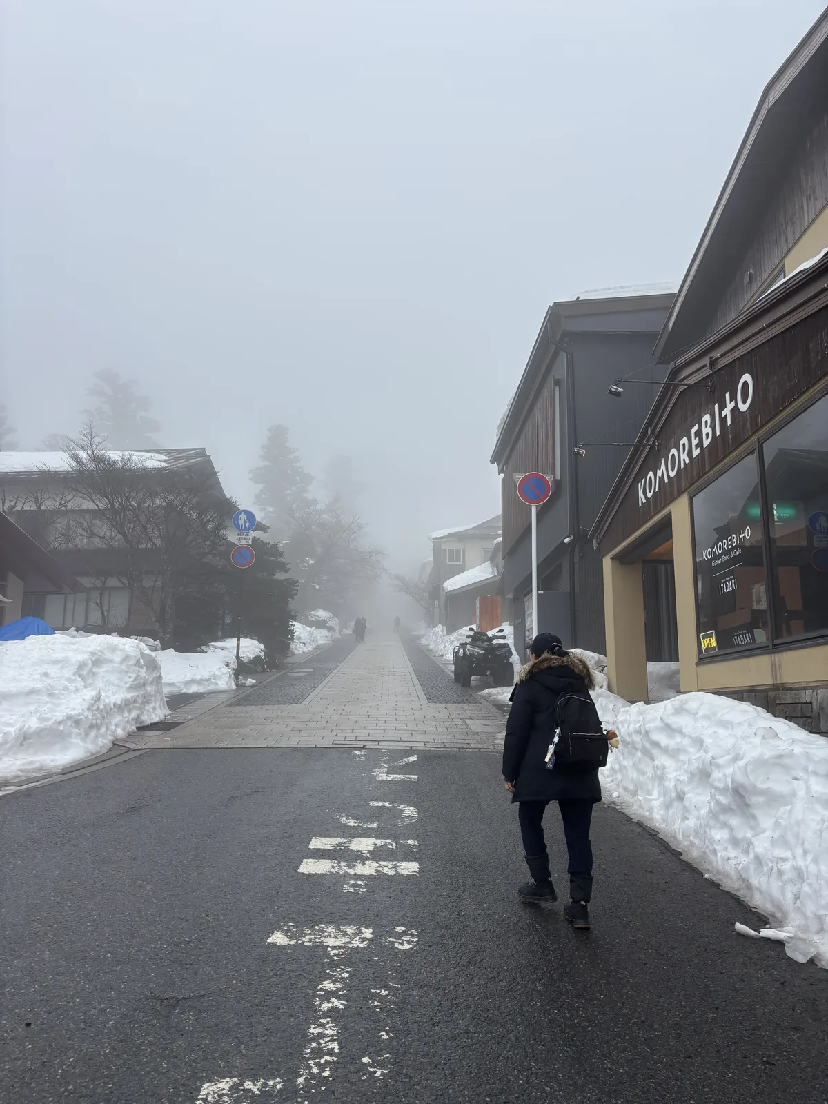 大山寺參道的入口一帶，右側是一棟深色外牆的商店，招牌上以英文寫著 KOMOREBITO，前方的道路覆蓋著薄雪，左側是堆高的積雪與另一棟建物，一位行人背向鏡頭走向霧中的參道
