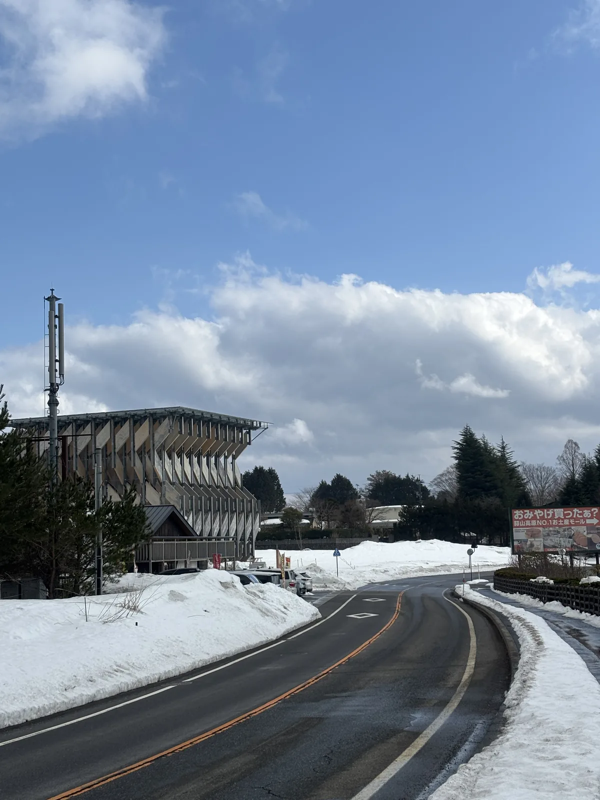 雪後的縣道向前方彎曲，兩側堆著積雪，左前方是木構展亭的側面，斜向的木板與上方出挑的屋頂清晰可見，展亭右側是一排針葉樹，天空有大片白雲