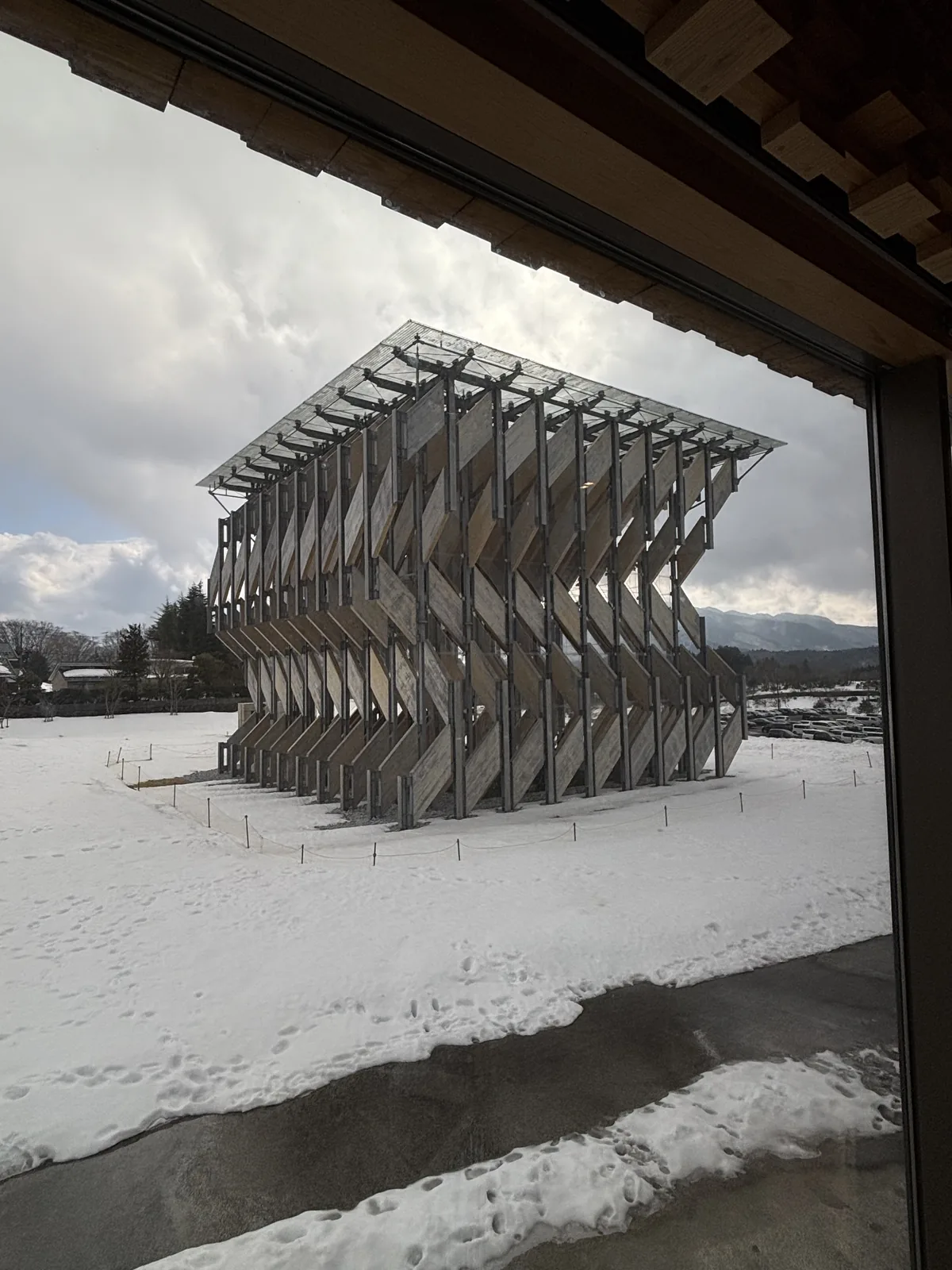 從美術館棟室內透過大片玻璃望出的風之葉，展亭立在覆雪的草地上，四面由斜向的木板構成，畫面上緣是室內木構的簷口與窗框