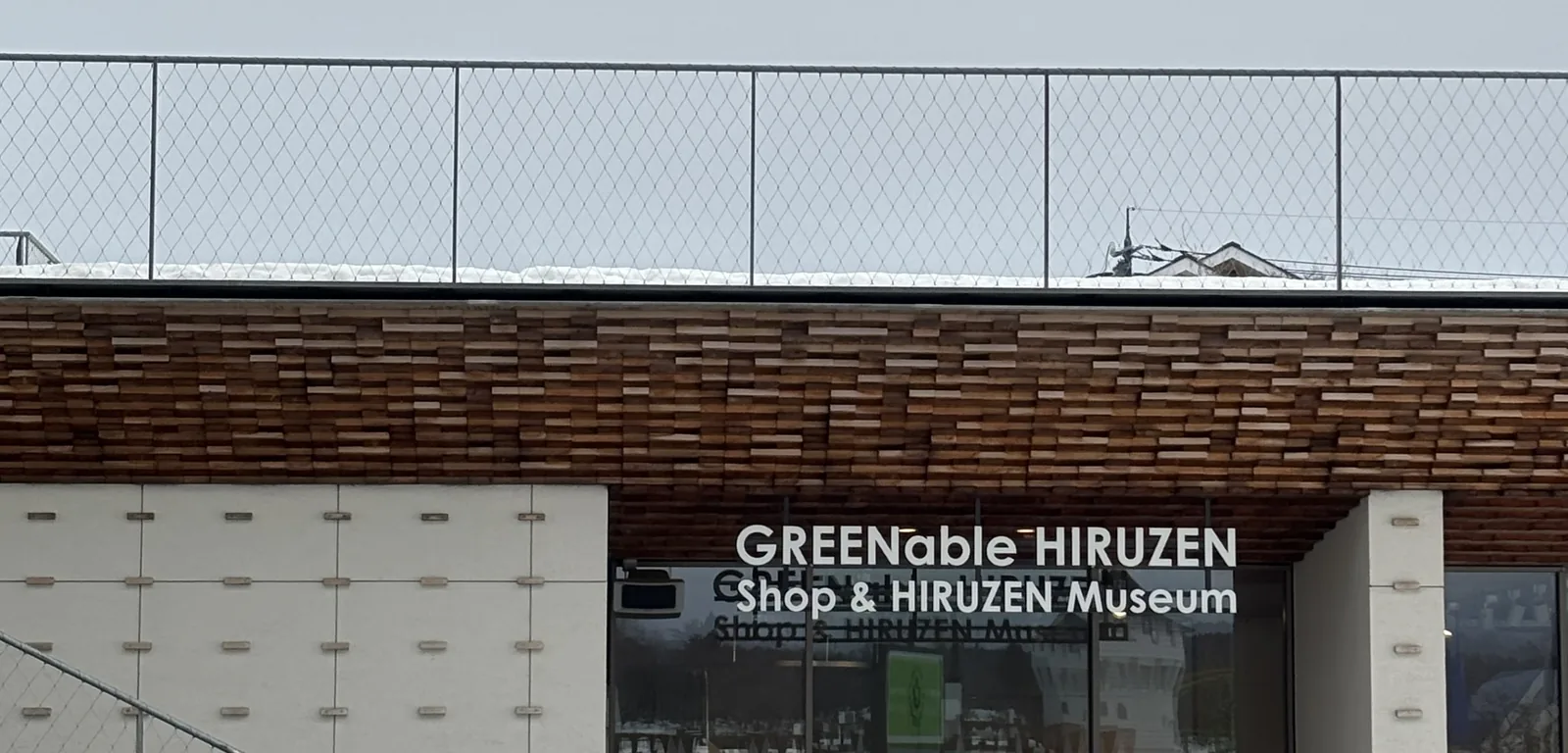 美術館棟的入口，簷口由深褐色的小木塊斷面層層排列成帶狀，下方左側是白色的牆板，右側是玻璃門，門上以英文寫著 GREENable HIRUZEN Shop & HIRUZEN Museum