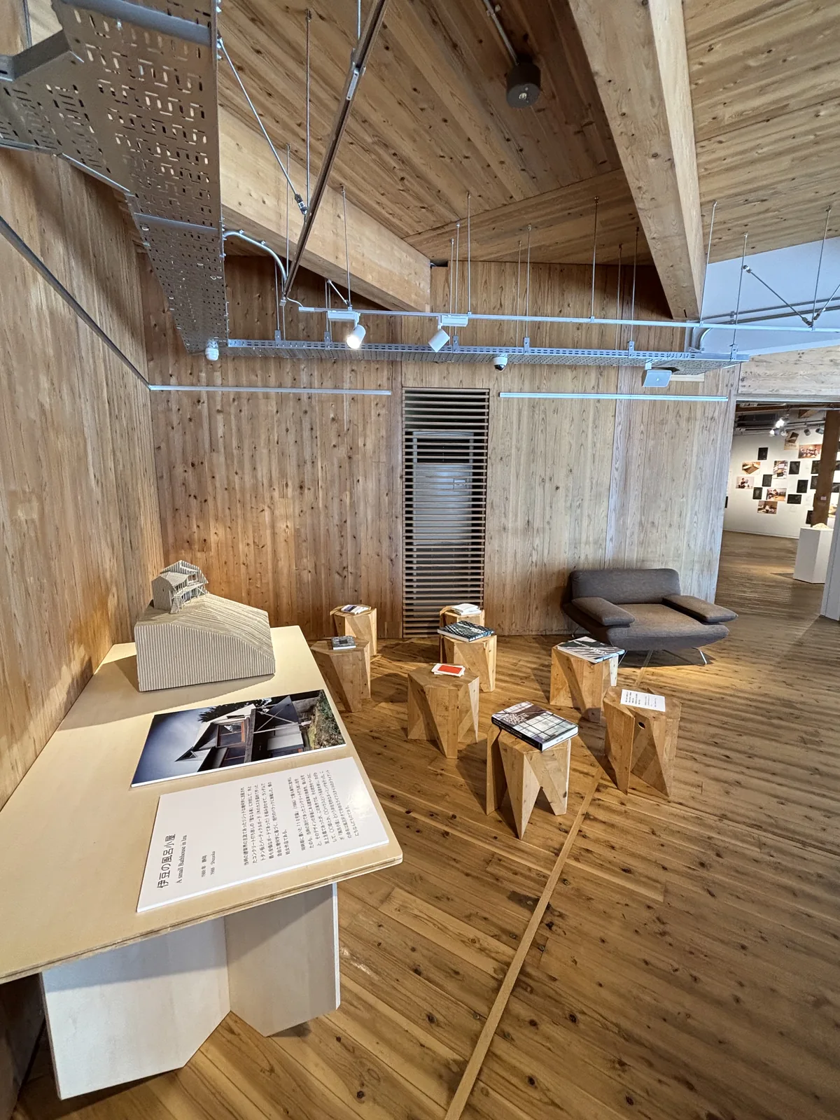 二樓展示廊，牆面與天花是大片的木板，天花一側沿斜度升起，室內有展示台、模型、數張多角形的木凳與一張灰色沙發，地面是木地板