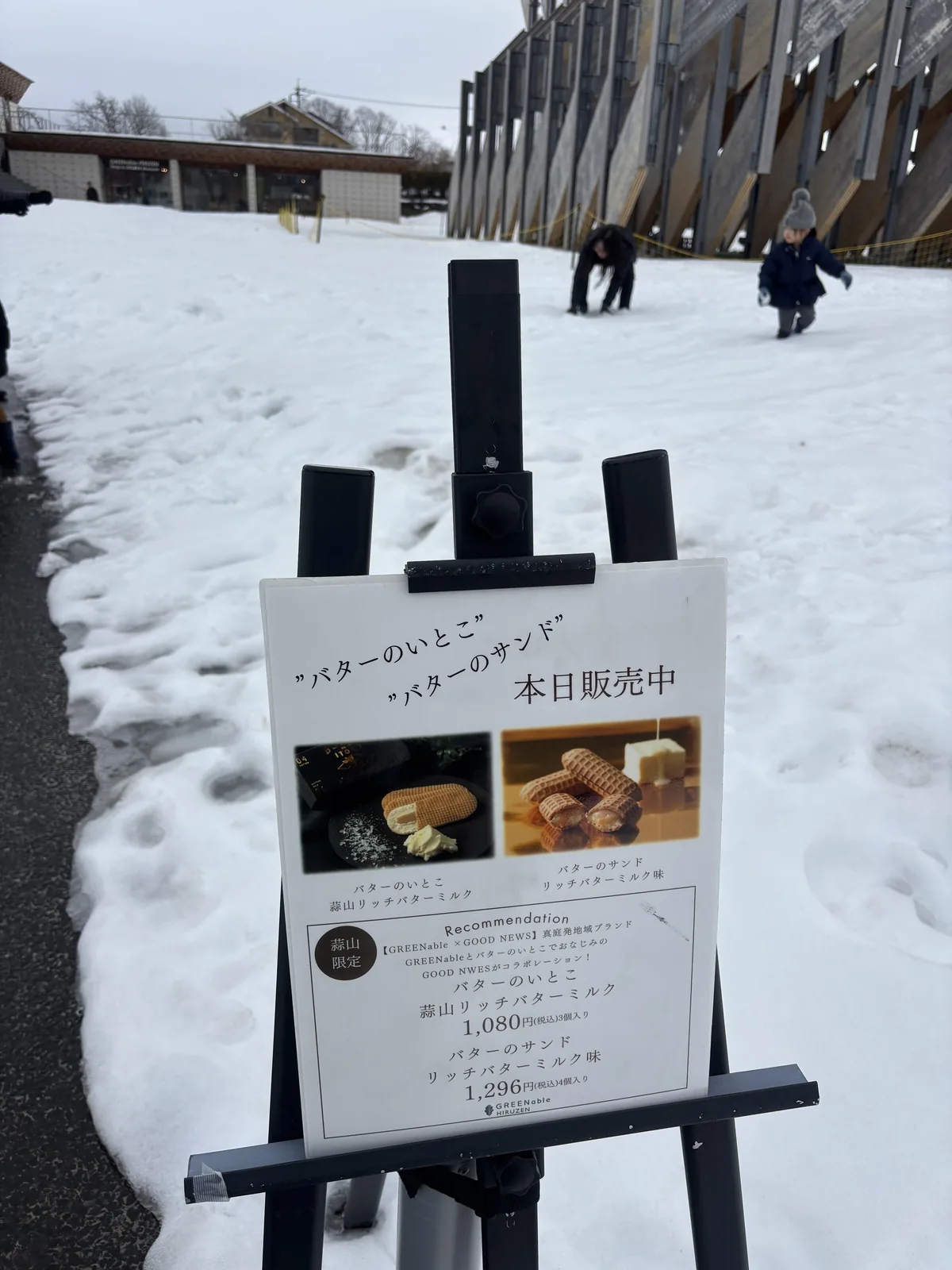 雪地上的一座黑色立架，上面夾著一張宣傳立牌，立牌上是兩款奶油夾心點心的照片與價格，背景是覆雪的斜坡與遠處的展亭