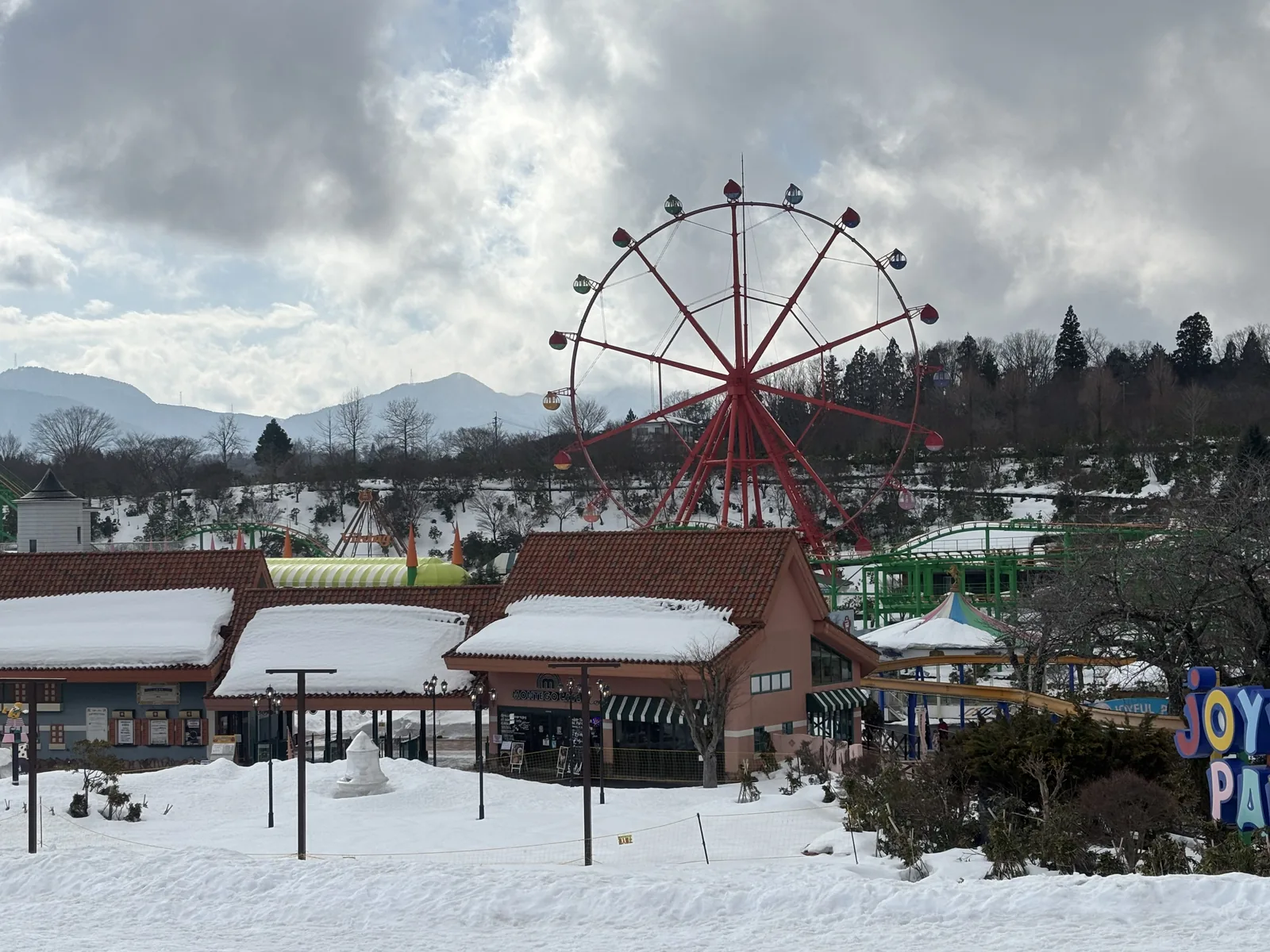 蒜山高原中心的遊樂園，前景是覆雪的草地，中景是一排紅色屋頂的商店建築，右後方是一座紅色的摩天輪，背景是覆雪的山與陰天