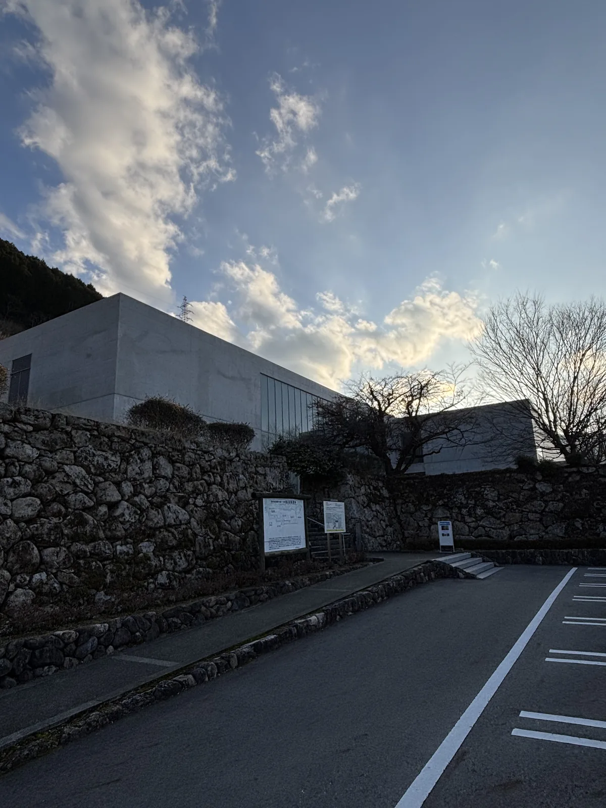 自停車場望美術館，前景是柏油的停車場與白色的停車格線，中景是一段以大小不一的石塊砌成的石垣，石垣上方是美術館的清水混凝土量體與玻璃面，右側是落葉的樹