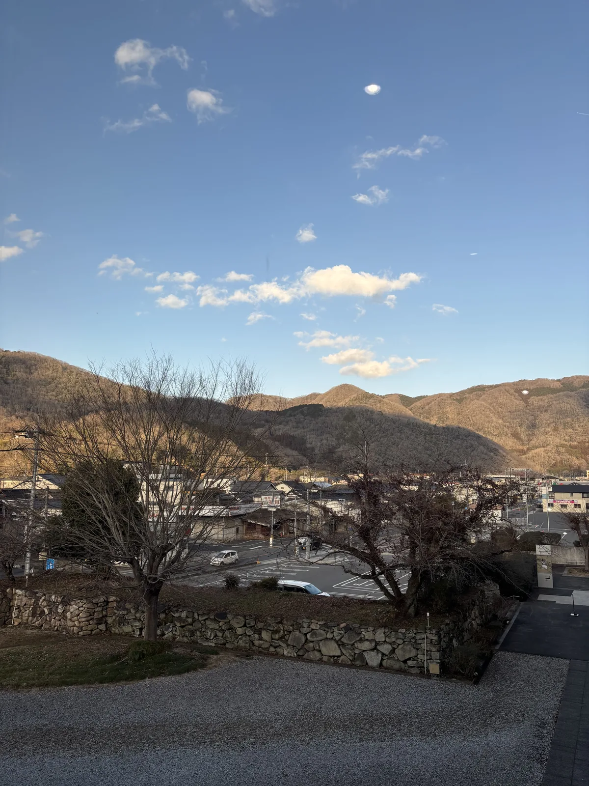 自美術館高處向北回望，前景是鋪著碎石的平台與石垣頂端，中景是落葉的樹、停車場與成羽的民家，背景是夕照下的山稜與藍天