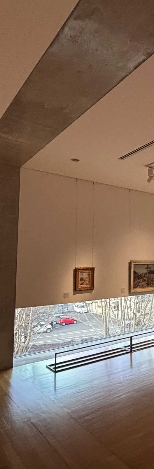 二樓展示室的一角，左側是一根斜向穿過天花的清水混凝土梁與混凝土柱，正面是白色的展示牆與兩幅畫作，牆下方有一道貼近地面的橫長窗，窗外是停車場與落葉的樹，地面是木地板