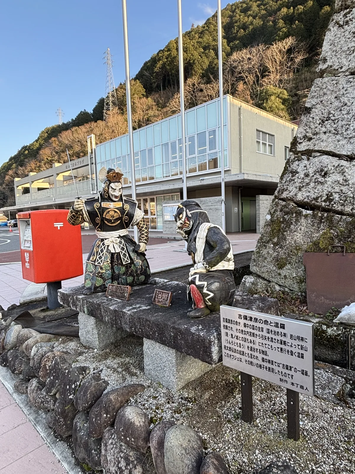 Taikomaru Plaza 前的廣場，中央是一座紅色郵筒與兩尊彩色人像坐在石凳上，旁邊立著一塊解說牌，後方是一棟兩層的白色建築與旗桿，右側是陣屋的石垣，背景是長滿樹的山