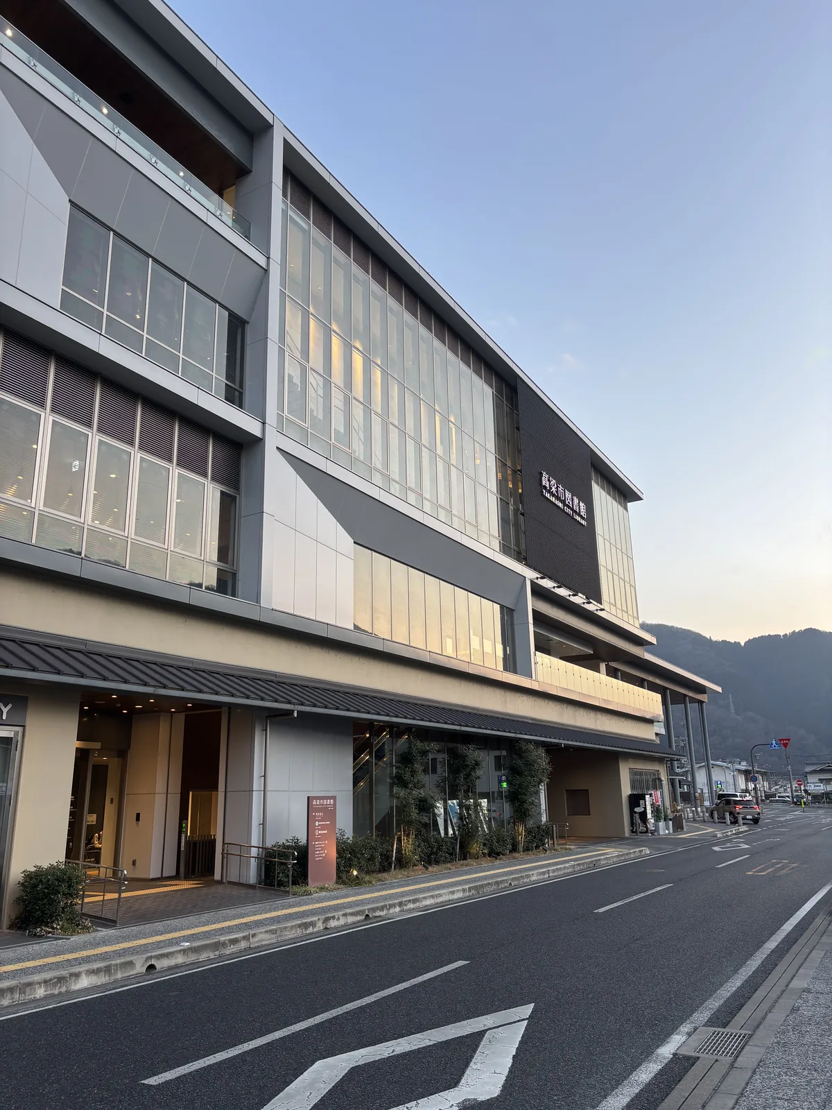 自站前道路望複合設施，左側是四層的建物，立面由白色與灰色的牆板、橫長的玻璃窗與直向的格柵構成，一樓上方有一道深色的出簷，右側遠處是暮色中的山，前景是空曠的車道