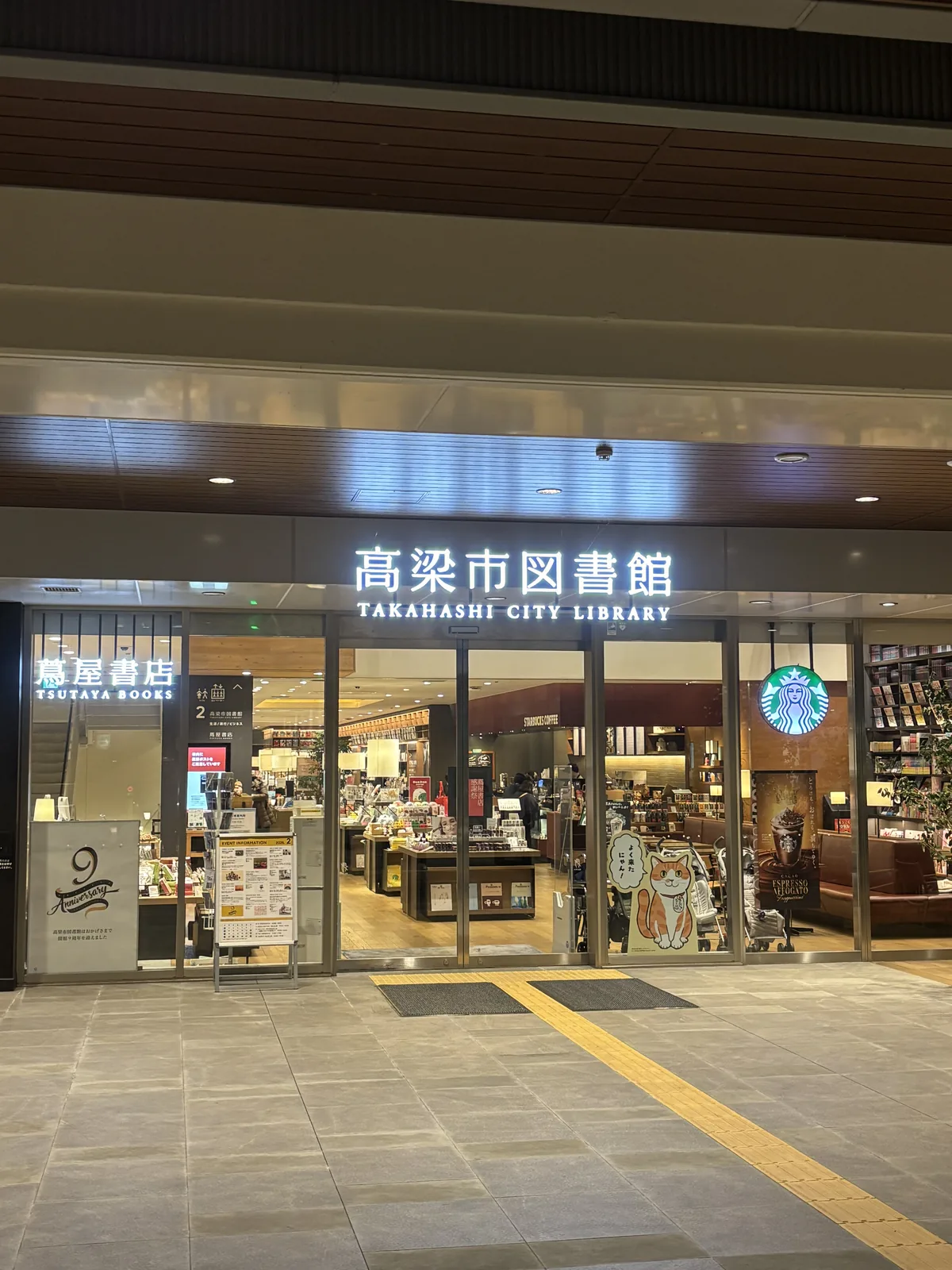 夜間的圖書館入口，上方是深色的木條天花與嵌燈，中央是寫著高梁市圖書館與英文館名的玻璃門，門內可見書架與商品陳列，左側掛著蔦屋書店的招牌，右側是 Starbucks 的標誌，地面是灰色石材與黃色導盲磚