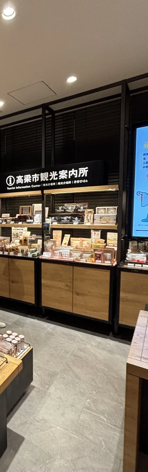 觀光案內所的櫃台，上方是寫著高梁市觀光案內所的黑底招牌，下方是木製的櫃台與層架，陳列著盒裝的土產與點心，左側是灰色的石材地面，右側是一面數位看板