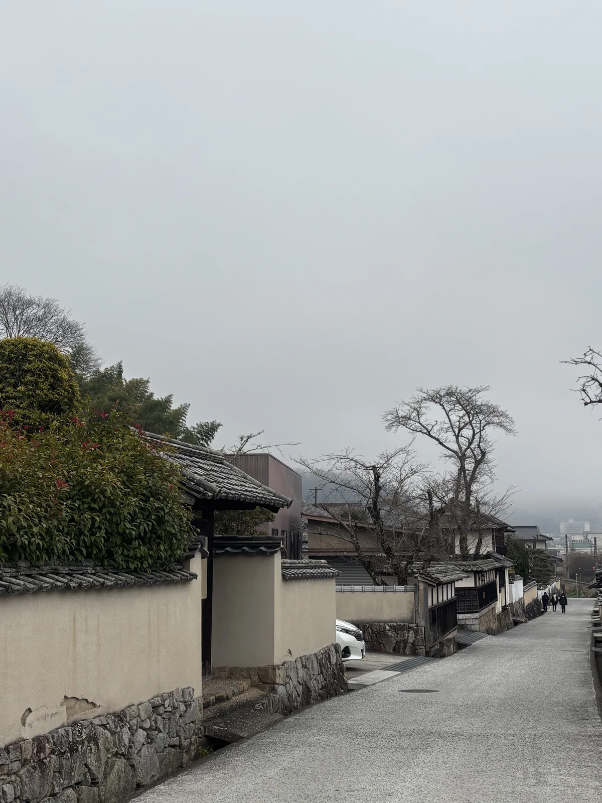 石火矢町的街道，左側是白色的土塀與石垣，土塀上方覆瓦，中央是一條筆直的石鋪道路，遠處是落葉的樹與霧中的山