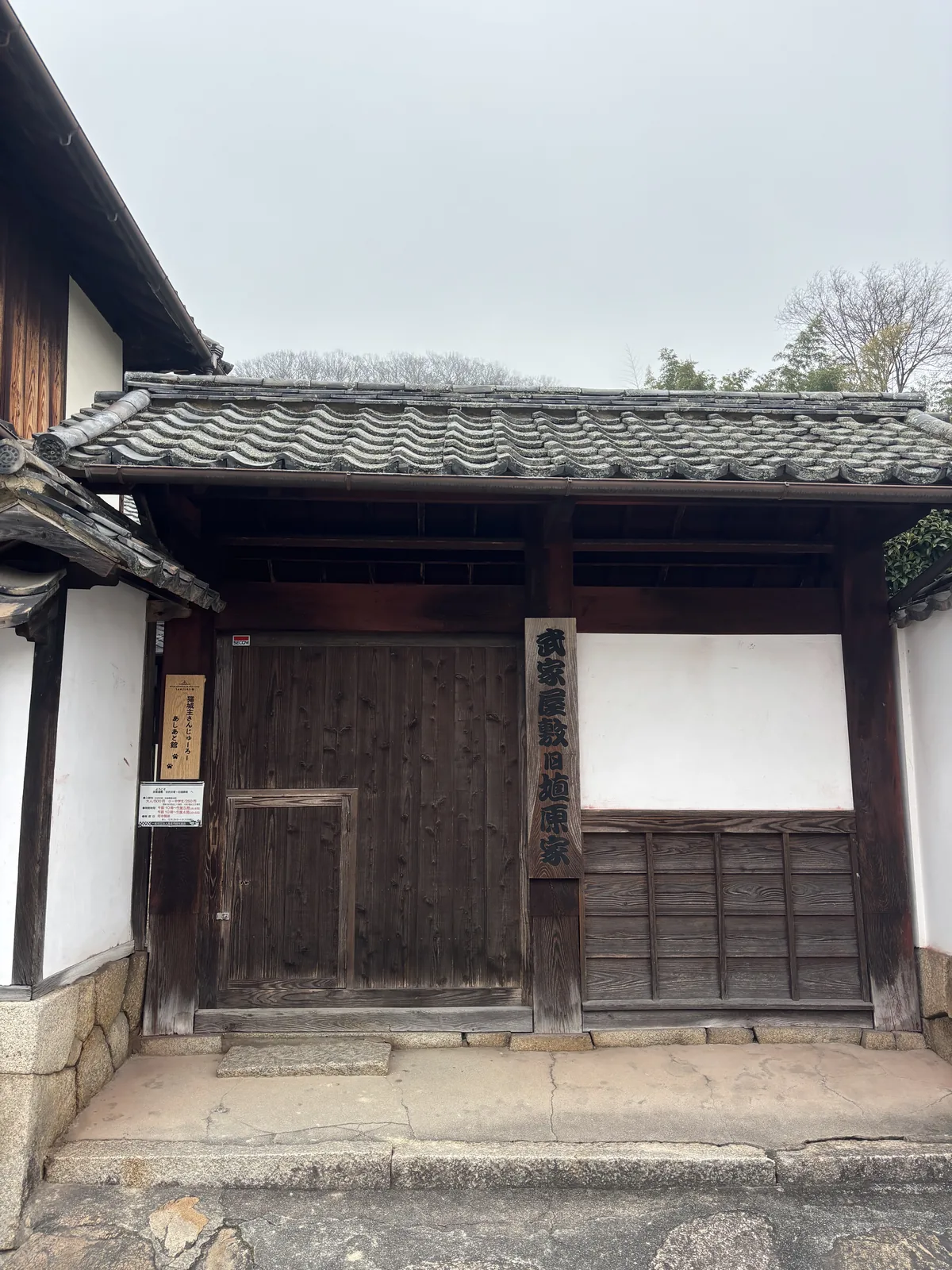 一座武家屋敷的門，木造的門扉與門柱呈深褐色，上方覆著瓦屋頂，兩側是白色的牆與石砌的基座，門旁掛著一塊木牌