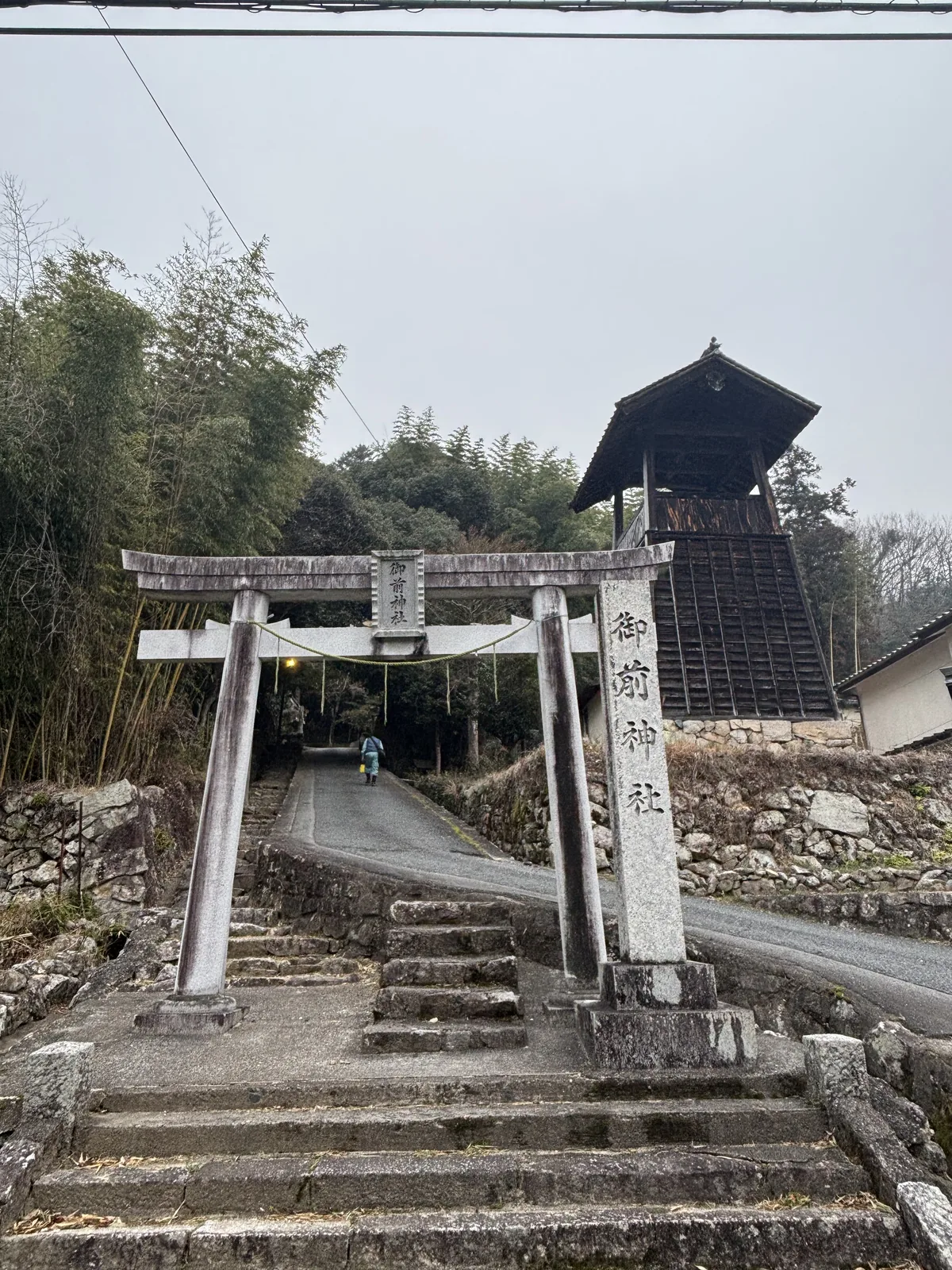 御前神社的石造鳥居，鳥居後方是一段石階與上坡的道路，右後方是一座深色木造的塔狀建物，周圍是竹林與石垣
