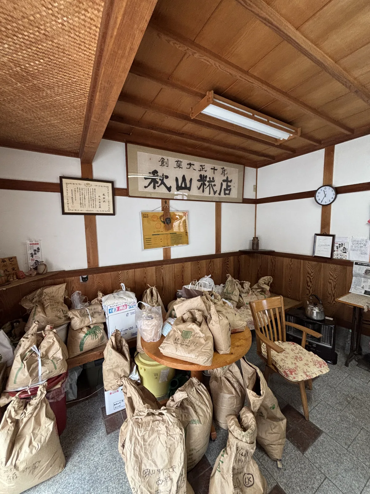 秋山糀店的店內，牆上掛著寫有店名的木製匾額與證書，下方是大量堆放的牛皮紙袋，中央是一張小圓桌與兩張椅子，天花是木構的格子