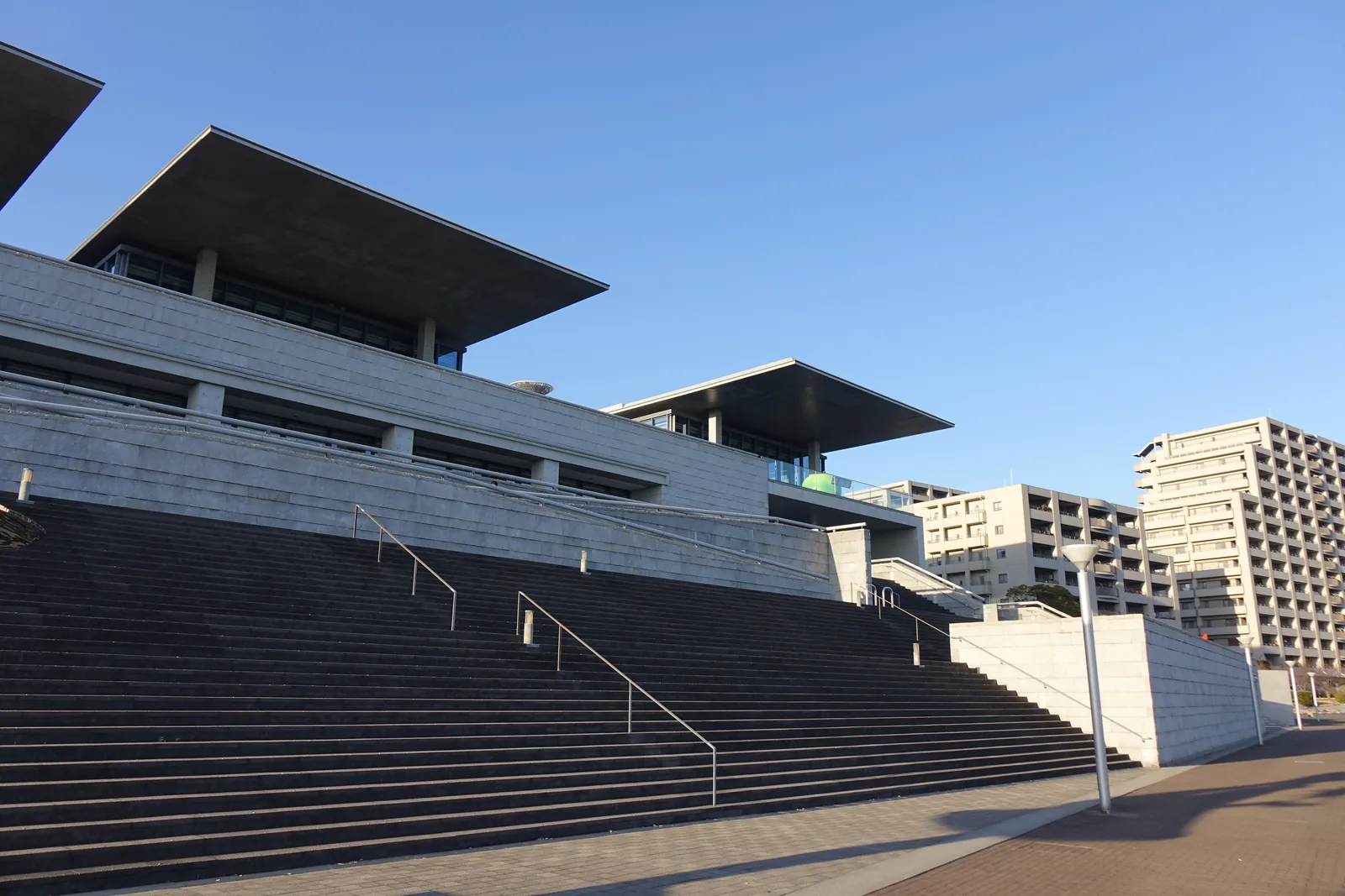 兵庫縣立美術館的南側外觀，前景是一段寬闊的石造大階梯，階梯之上是並排的量體，每一棟的頂部以薄而深的混凝土屋簷向外挑出，屋簷下是玻璃立面，右側遠處可見一顆綠色的蘋果與住宅大樓