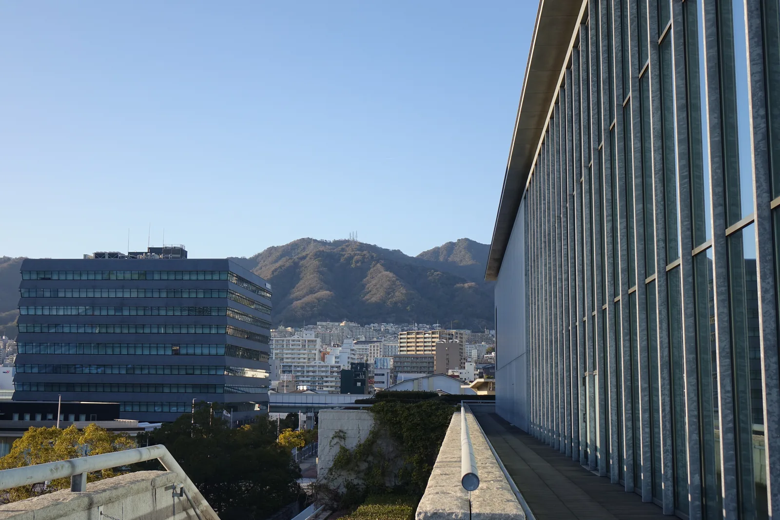 自美術館高處望向北方，左側是高架道路與辦公大樓，中央遠處是六甲山系的山稜，右側是美術館的玻璃立面與石造的矮牆