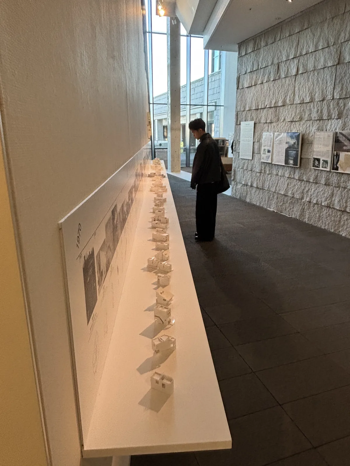Ando Gallery 內的一側，左側是白牆前一道長長的白色展示台，台上排列著一列白色的建築模型，右側是割肌的天然石貼牆，遠處是玻璃面與一位來館者