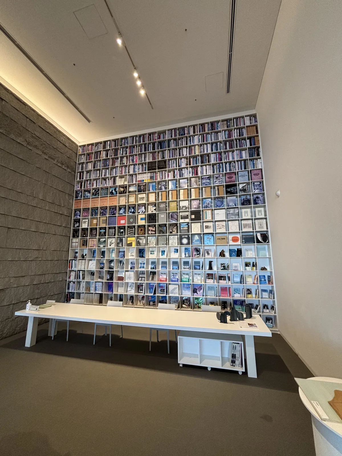 Ando Gallery 上層的 library corner，正面是一面從地面到天花的書架，書架上整齊排列著書，前方是一張白色的長桌，左側是割肌的石牆，天花有線狀的照明