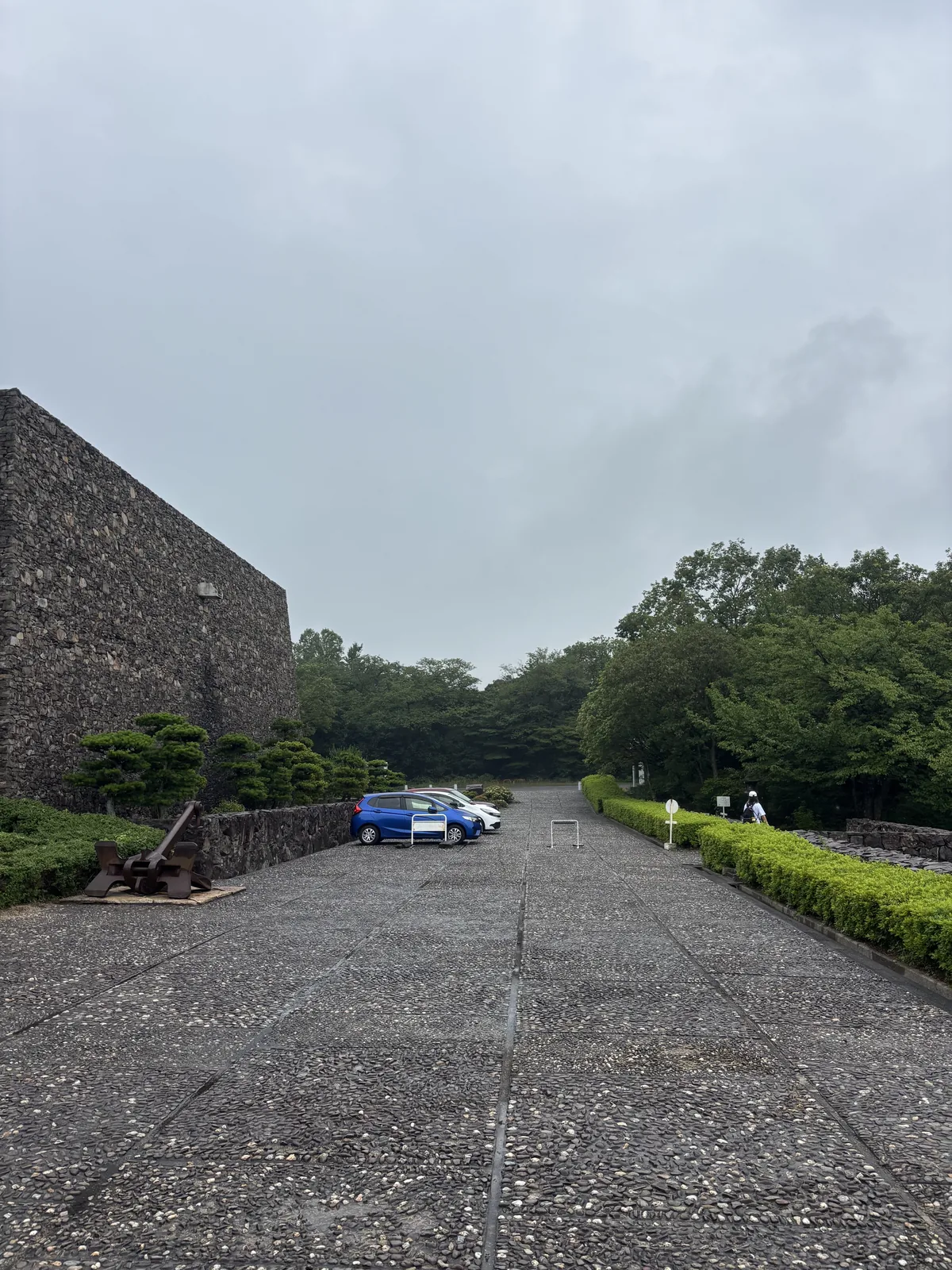 自停車場望資料館，左側是一座無窗的石積量體，中央是一條鋪著碎石的寬闊通道，兩側是修剪整齊的綠籬，遠處停著兩輛車，背景是樹林與陰天
