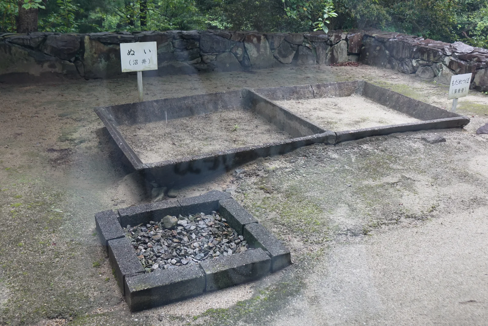 隔著玻璃窗所見的屋外展示，地面是砂土，中央是以混凝土塊圍成的兩個淺池與一個裝著碎貝殼的方形槽，旁邊立著一塊白色的小牌，背後是石積的擋土牆與樹林