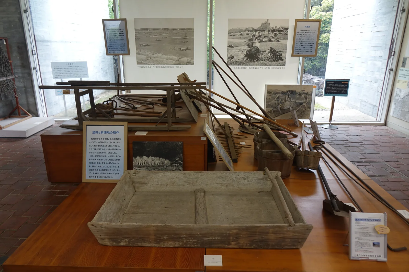 鹽田用具的展示室，前景是木製的展示台與一個淺木箱及耙具，後方兩側是整面落地玻璃，中間是一段清水混凝土牆，牆上掛著鹽田的黑白照片與解說，窗外是綠地